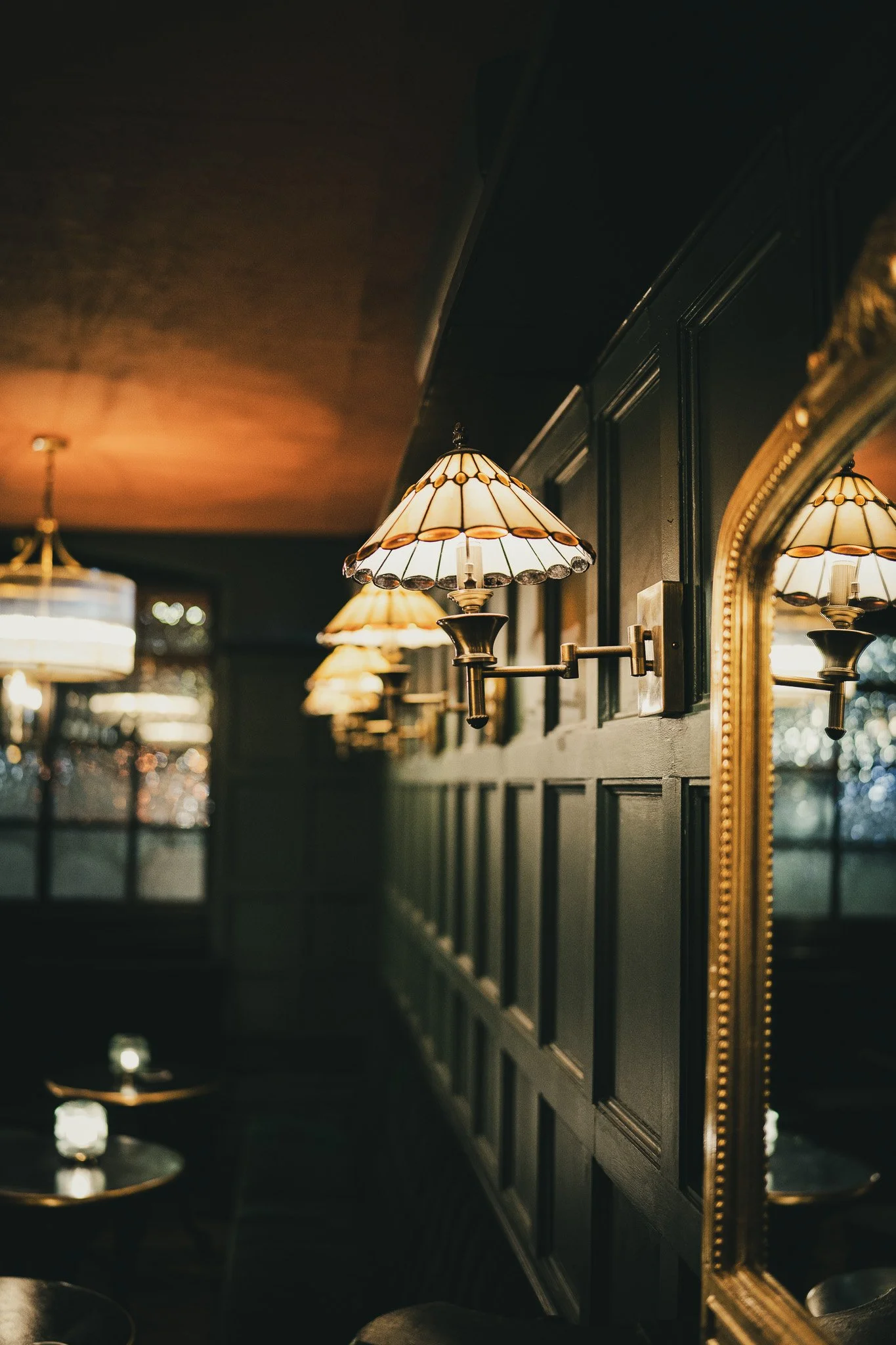 Inside a dimly lit restaurant or bar with decorative wall-mounted lamps with stained glass shades, a large ornate mirror, and wooden wall paneling.