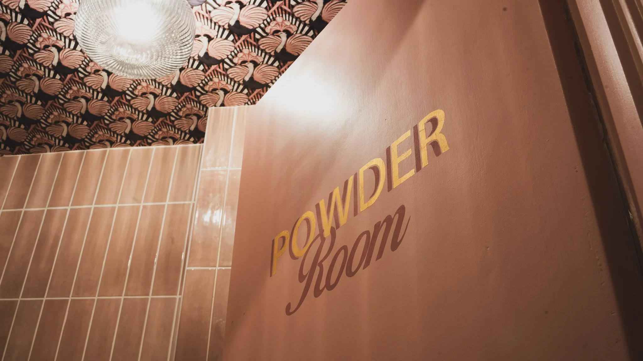 Close-up of a wall with the words 'Powder Room' in gold and brown letters. The wall is painted in a warm beige color. To the left, there are brown tiles, and in the background, a ceiling with flamingo-patterned wallpaper and a glass light fixture.