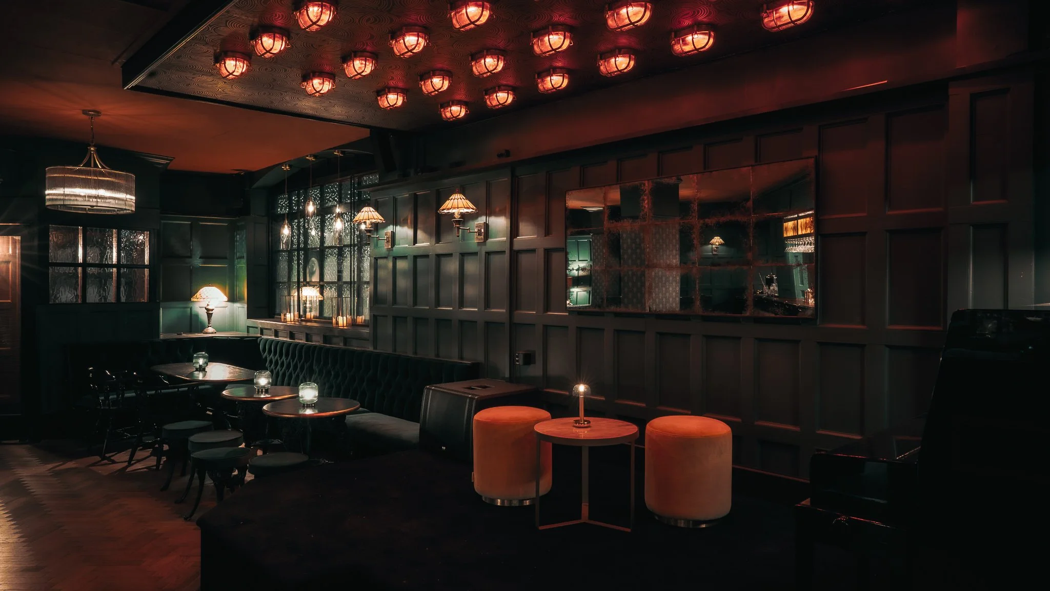 Dimly lit upscale bar or lounge with dark wood paneling, ambient hanging lamps, and a velvet booth seating area with small tables and glowing candles.