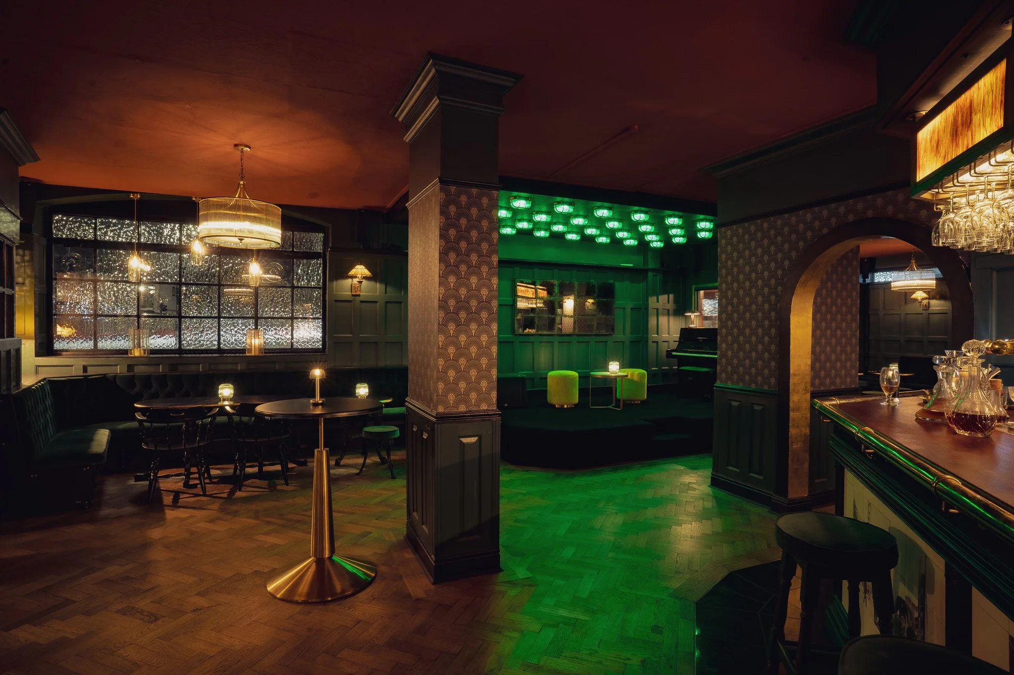 Interior of a dimly lit bar or lounge with wooden floors, green upholstered chairs, a stage with green lighting, and decorative wall panels.