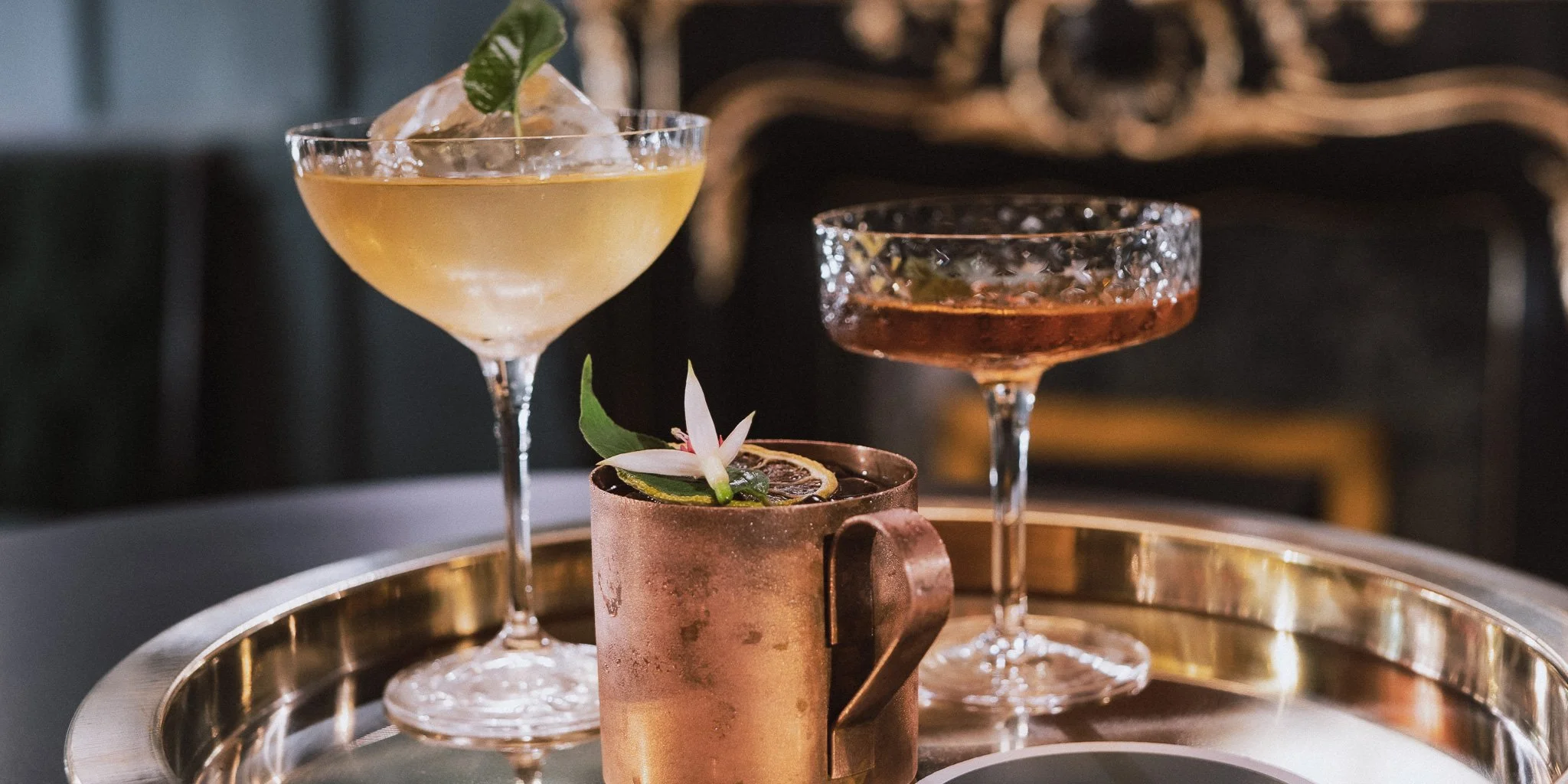 Three cocktails on a silver tray: a pale yellow cocktail with ice and a mint garnish in a coupe glass, a dark amber cocktail with ice in a coupe glass, and a dark drink with ice, garnished with a flower and citrus slice in a copper mug.