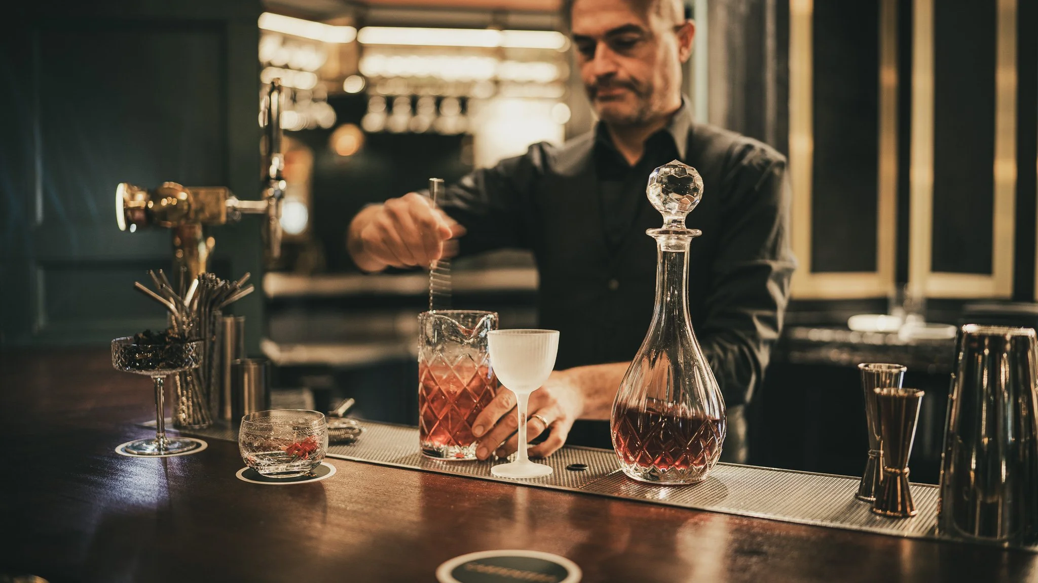 Bartender pouring a cocktail at a bar with various glassware and bar tools.