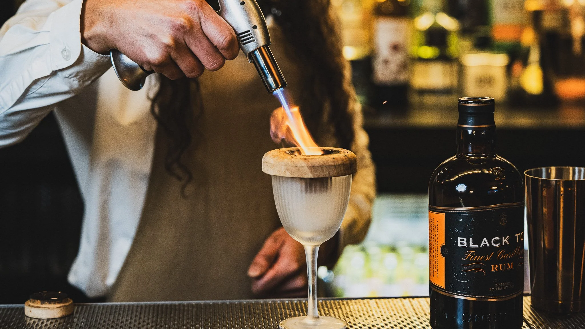A person using a blowtorch to add flame to a cocktail glass with a wooden and ceramic garnish on top, a bottle of Black Tot rum and a metal shaker nearby in a bar setting.