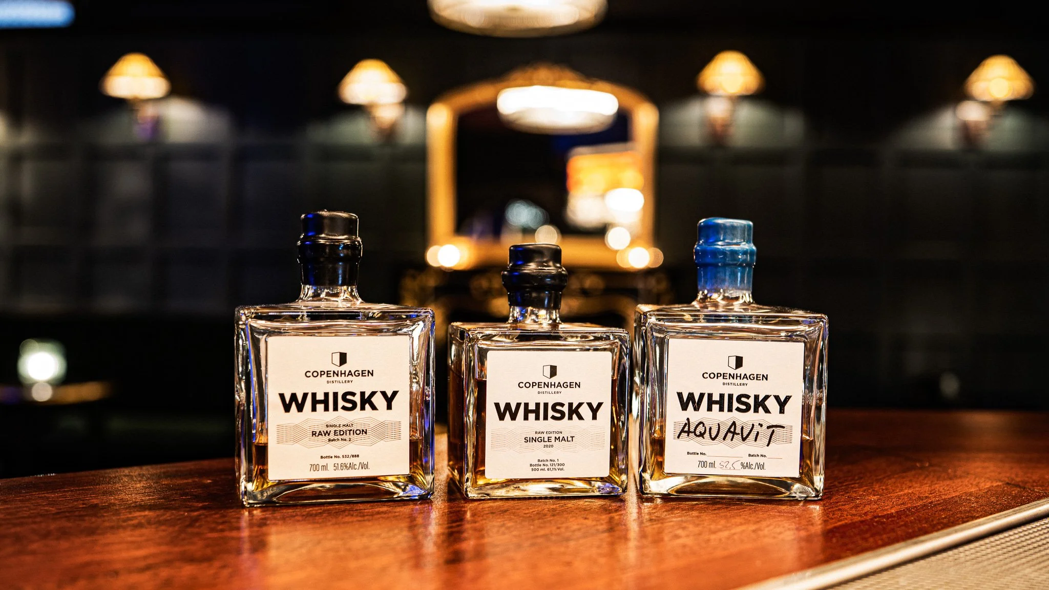 Three bottles of whiskey on a wooden bar counter in a dimly lit setting
