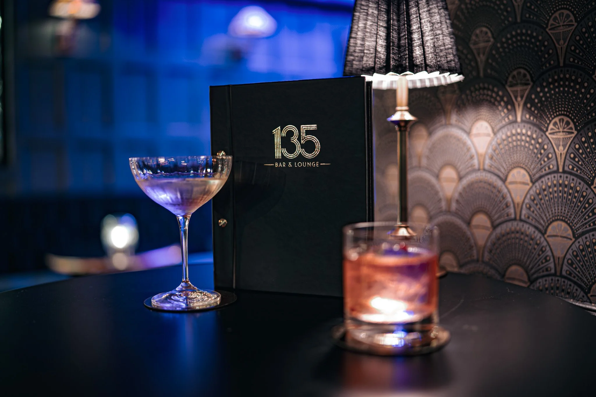 A black menu with gold text that reads '135 Bar & Lounge' on a table. There are two cocktails, one in a coupe glass and another in a short tumbler, with a lamp and a wall with decorative wallpaper in the background.