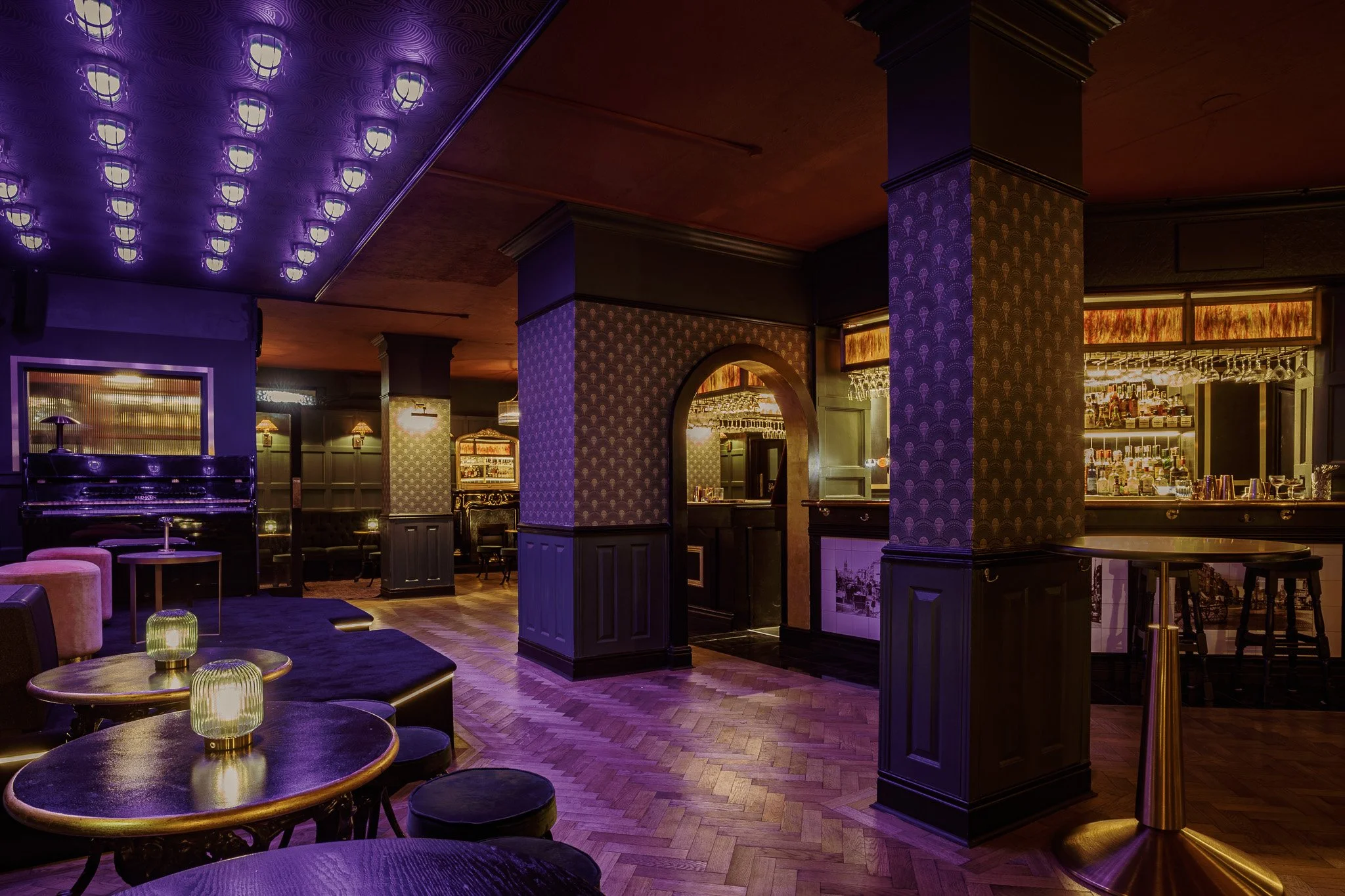 Interior of a bar or lounge with purple lighting, wooden floors, and dark wooden furniture. Bar area with bottles visible behind the counter, seating areas with tables and stools, and a decorative patterned wall.