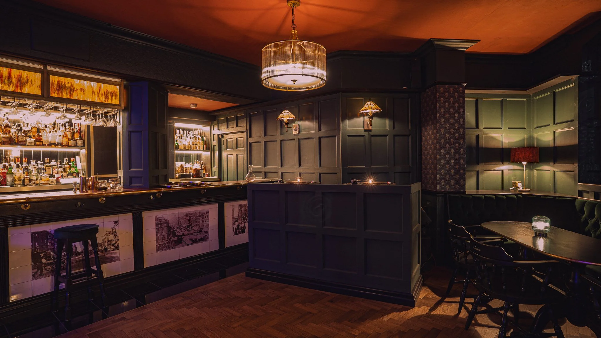 Inside a dimly lit bar with dark wood furnishings. There is a bar counter with various bottles of liquor and glasses, illuminated from behind. A round ceiling light and wall sconces light the space, and a seating area with a table, chairs, and a lamp