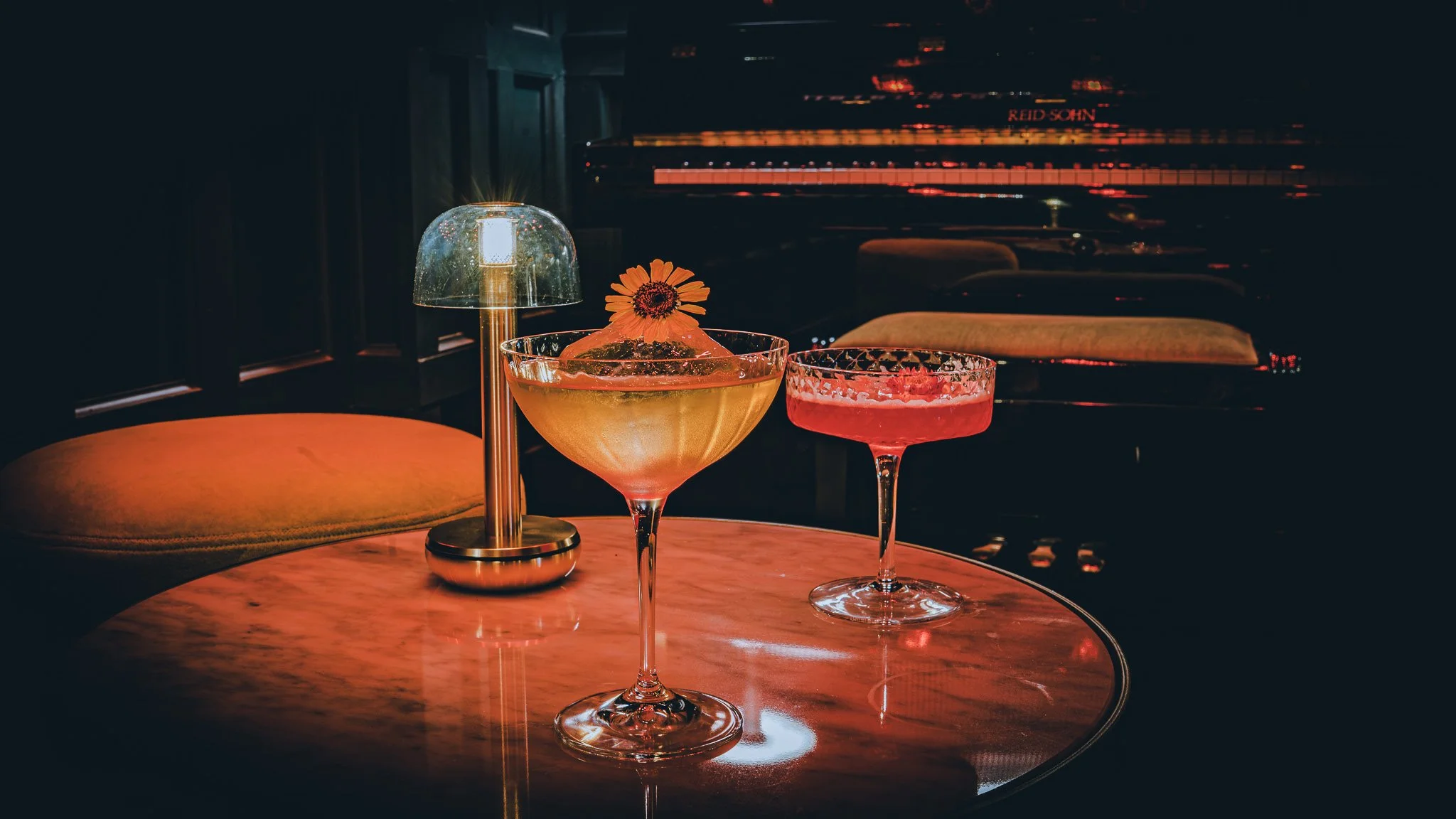 Two cocktail glasses and a small table lamp on a round wooden table in a dimly lit lounge with a piano in the background.