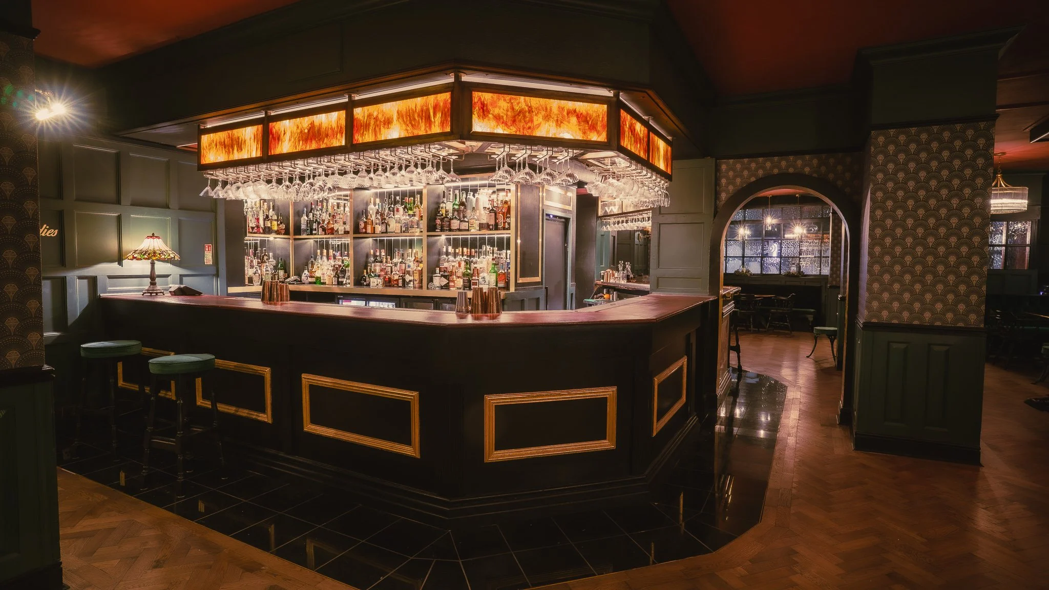Dimly lit bar with dark wood paneling, a curved black bar counter with gold accents, and a well-stocked liquor shelf. Hanging glasses are above the bar, and a warm, orange-toned light fixture decorates the ceiling. There are chairs and tables in the