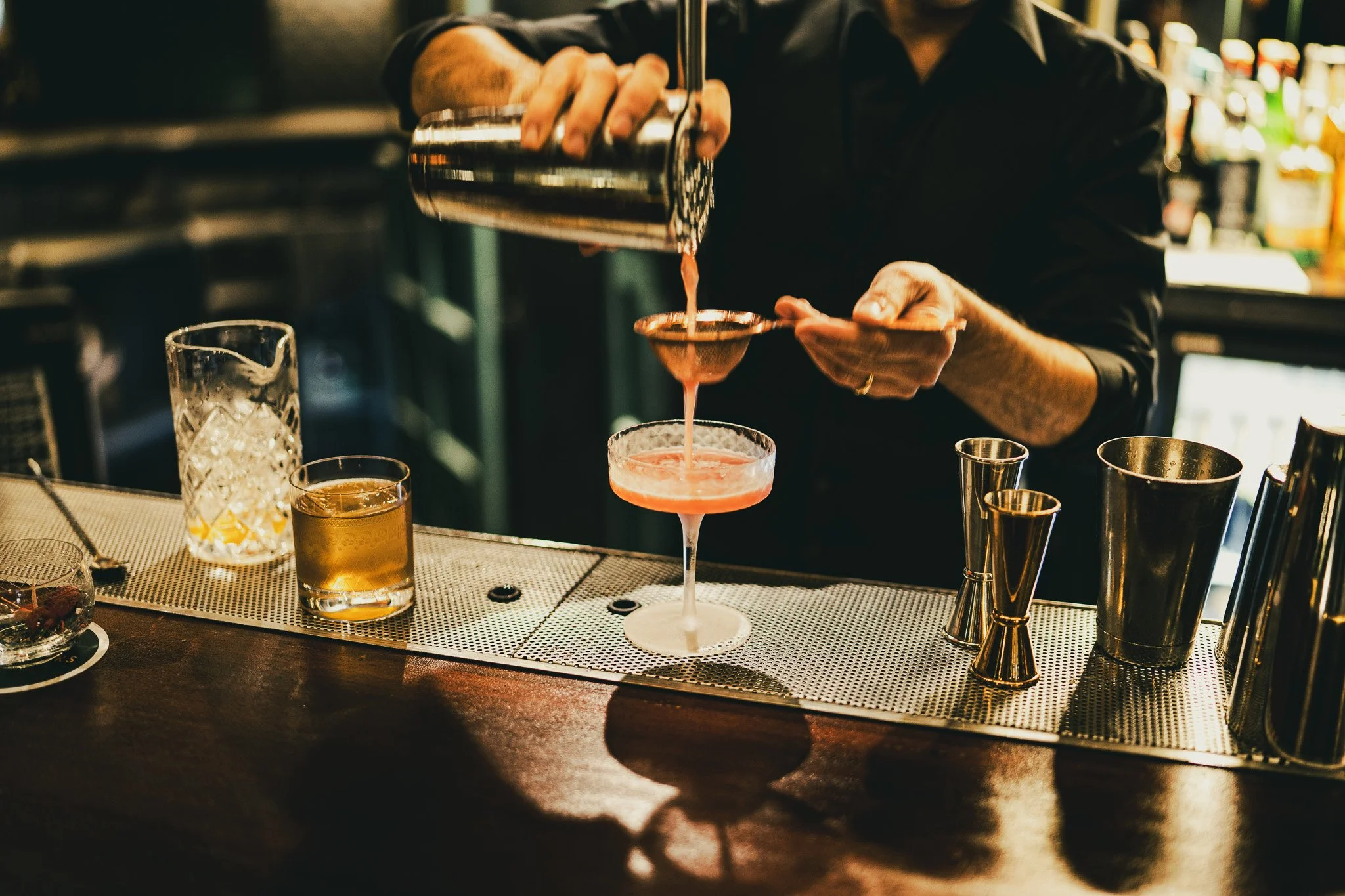 A bartender preparing a cocktail at a bar, pouring liquid through a strainer into a pink cocktail glass.