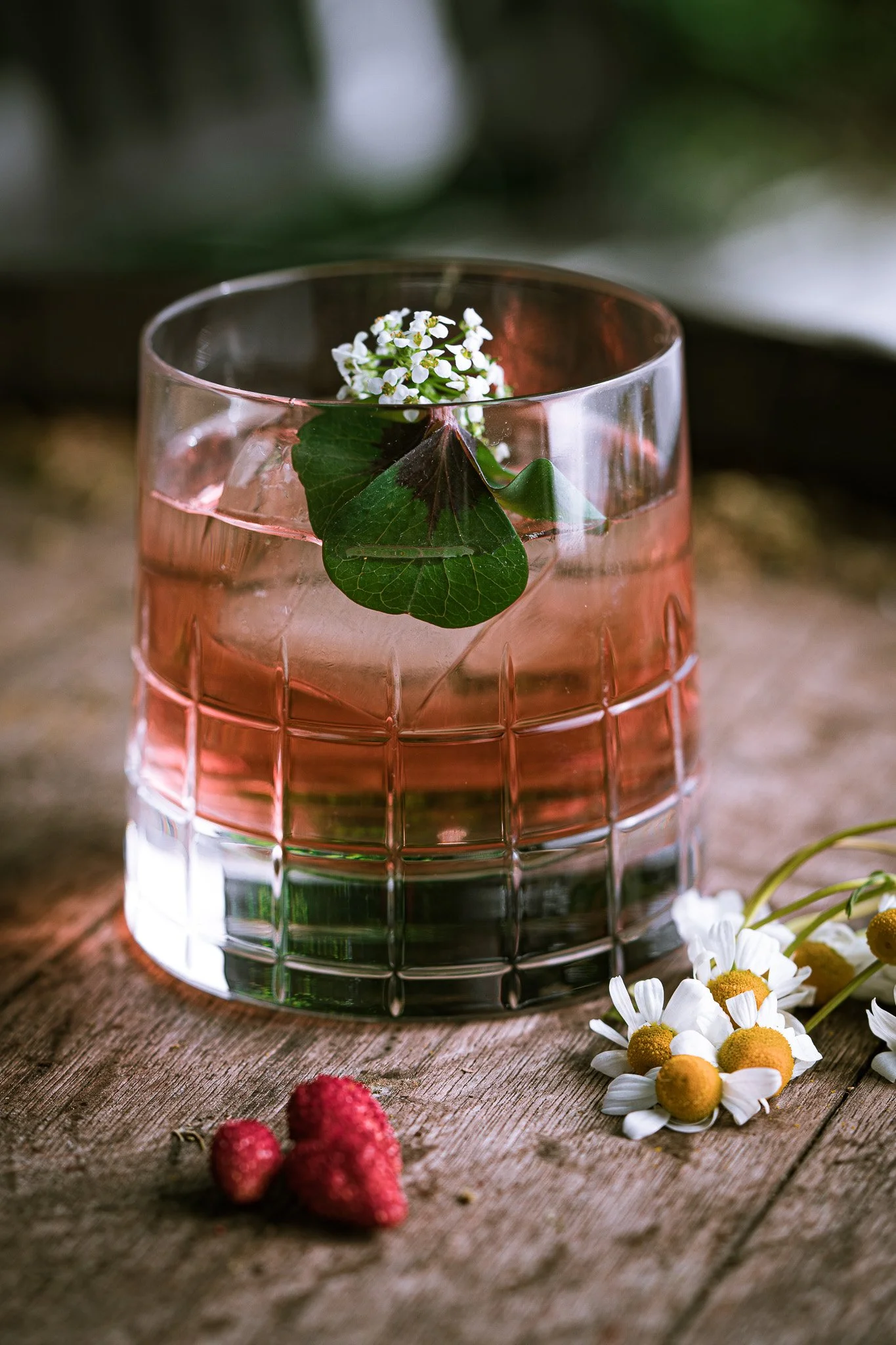 A glass with pink liquid, garnished with green ivy leaves and small white flowers, on a wooden surface with strawberries and white daisies nearby.