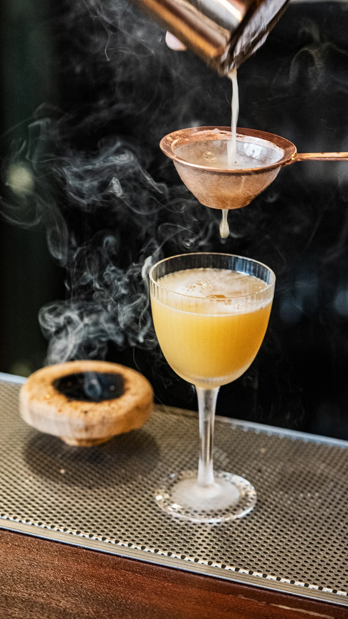 A cocktail glass filled with a yellowish drink, a wooden ring with a small fire, a steaming coffee dripper, and a coffee filter with a hot beverage, all on a metal surface with smoke or steam swirling around.