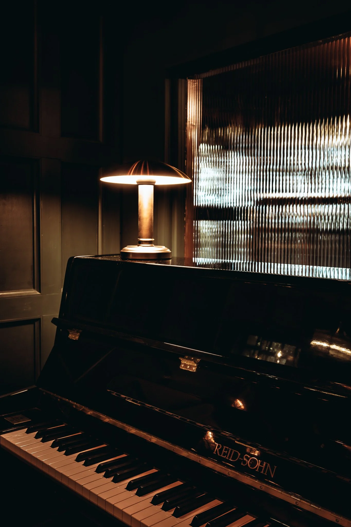 A darkly lit room with a black Reid-Sohn piano and a small table lamp emitting warm light, situated next to a window with vertical blinds.