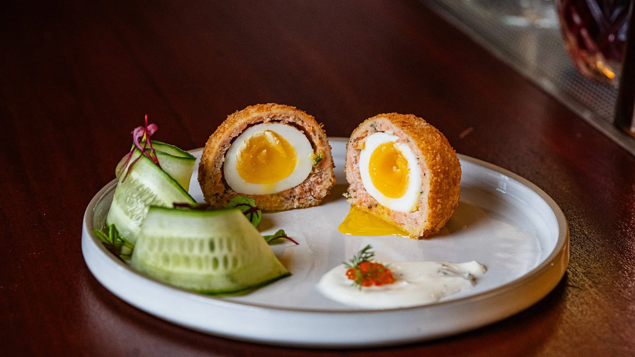 Deviled eggs with crispy breading, served with thinly sliced cucumber and garnished with microgreens and a dollop of sauce on a white plate.