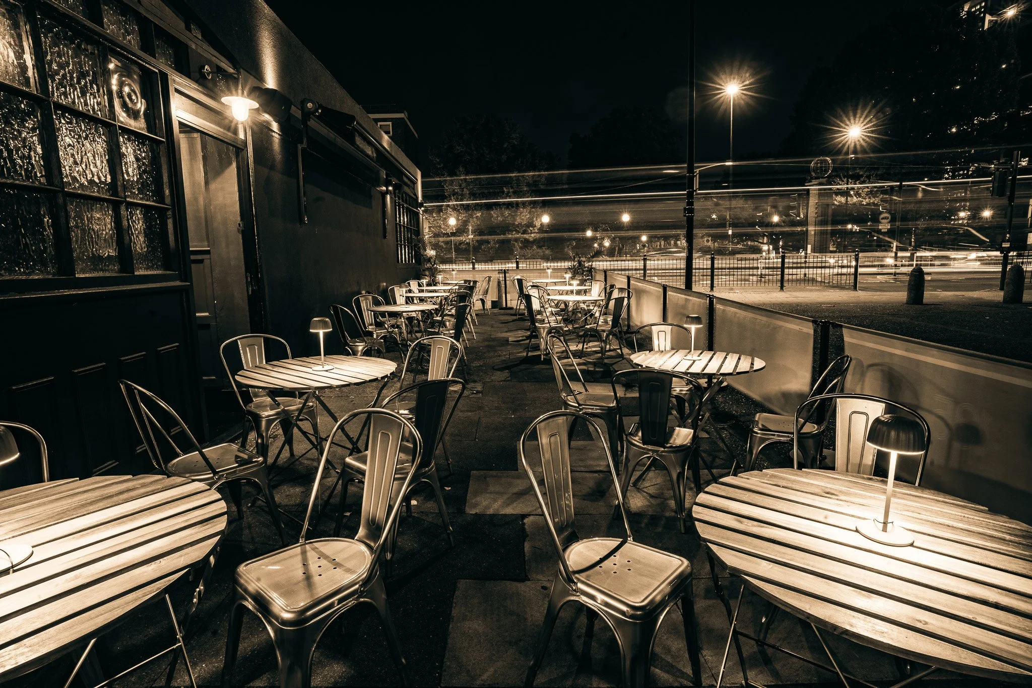 Outdoor seating area of a restaurant or cafe at night with empty round and rectangular tables, metal chairs, small lamps on tables, and a dark fence separating the patio from the street.
