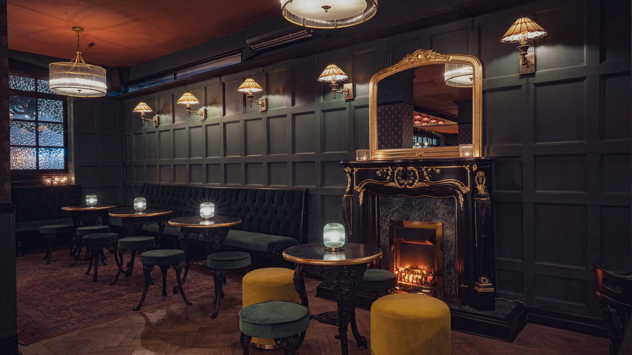 An elegant lounge with dark paneled walls, a fireplace with a gold-framed mirror above, and small glowing lamps on ornate wooden tables. There are green and yellow stools, tufted seating, and ambient lighting.