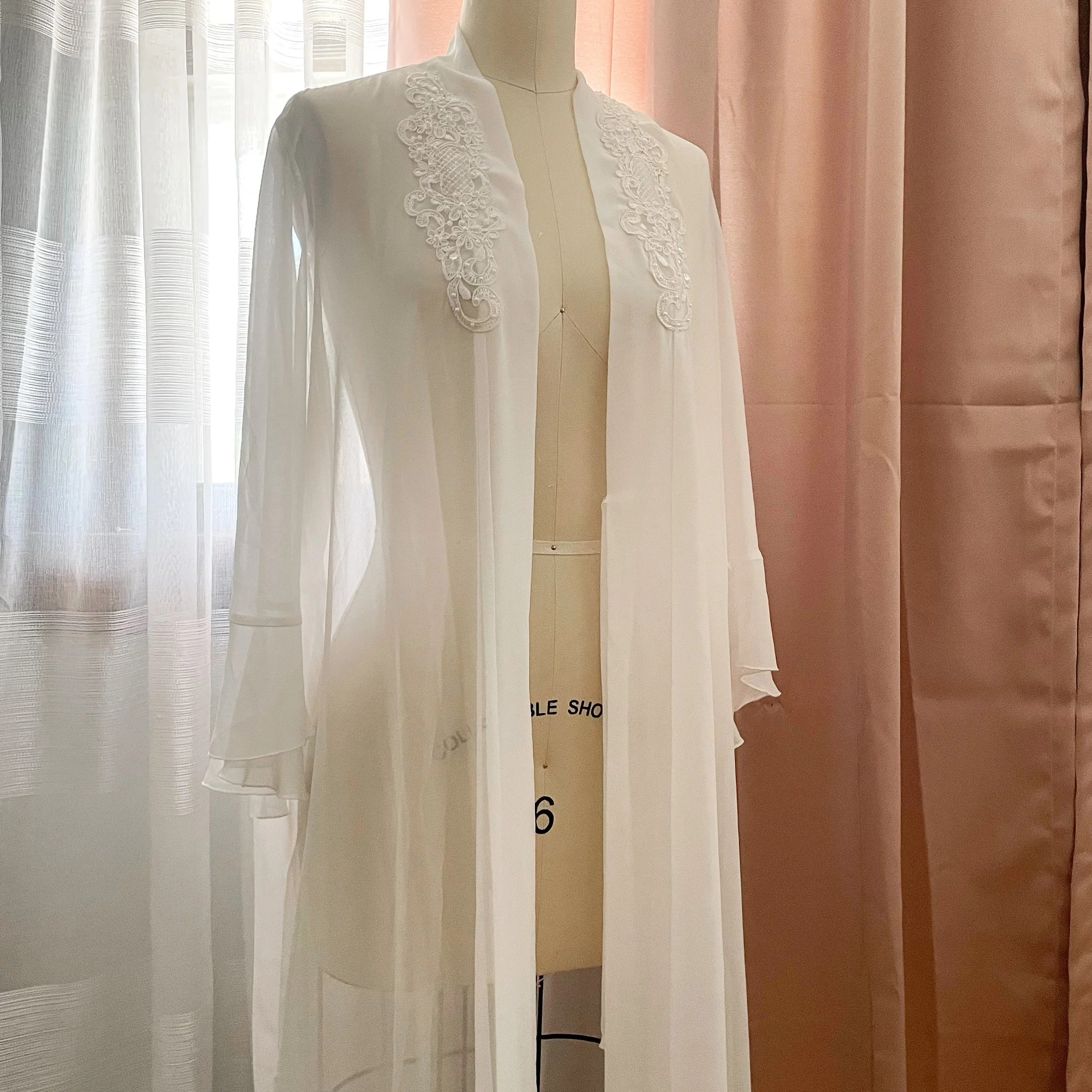White sheer robe with lace detailing on a dress form, set against a backdrop of sheer and solid curtains.