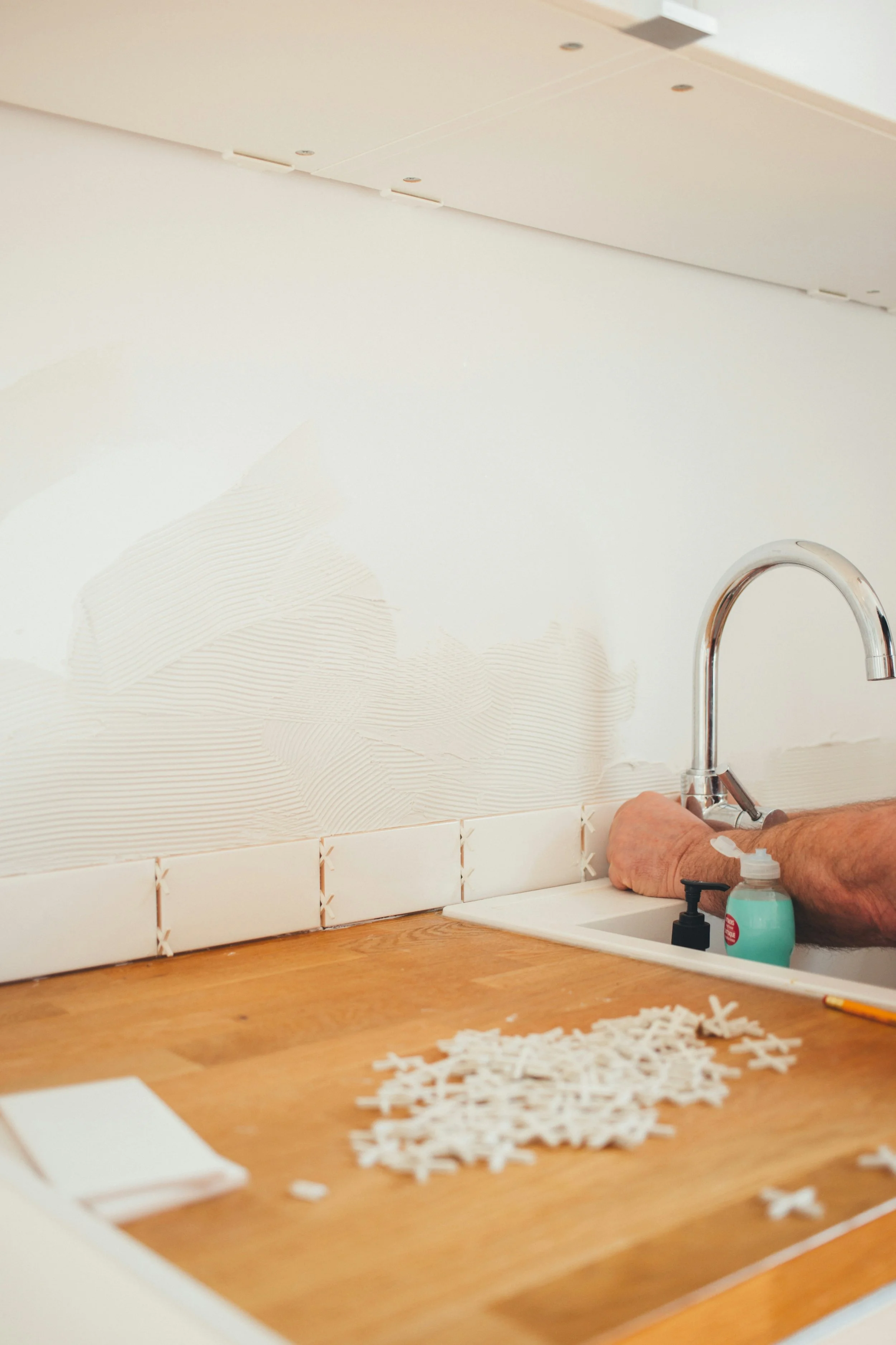 A person tiling a kitchen backsplash with white tiles; tile spacers are visible on a wooden countertop near a sink with a chrome faucet.