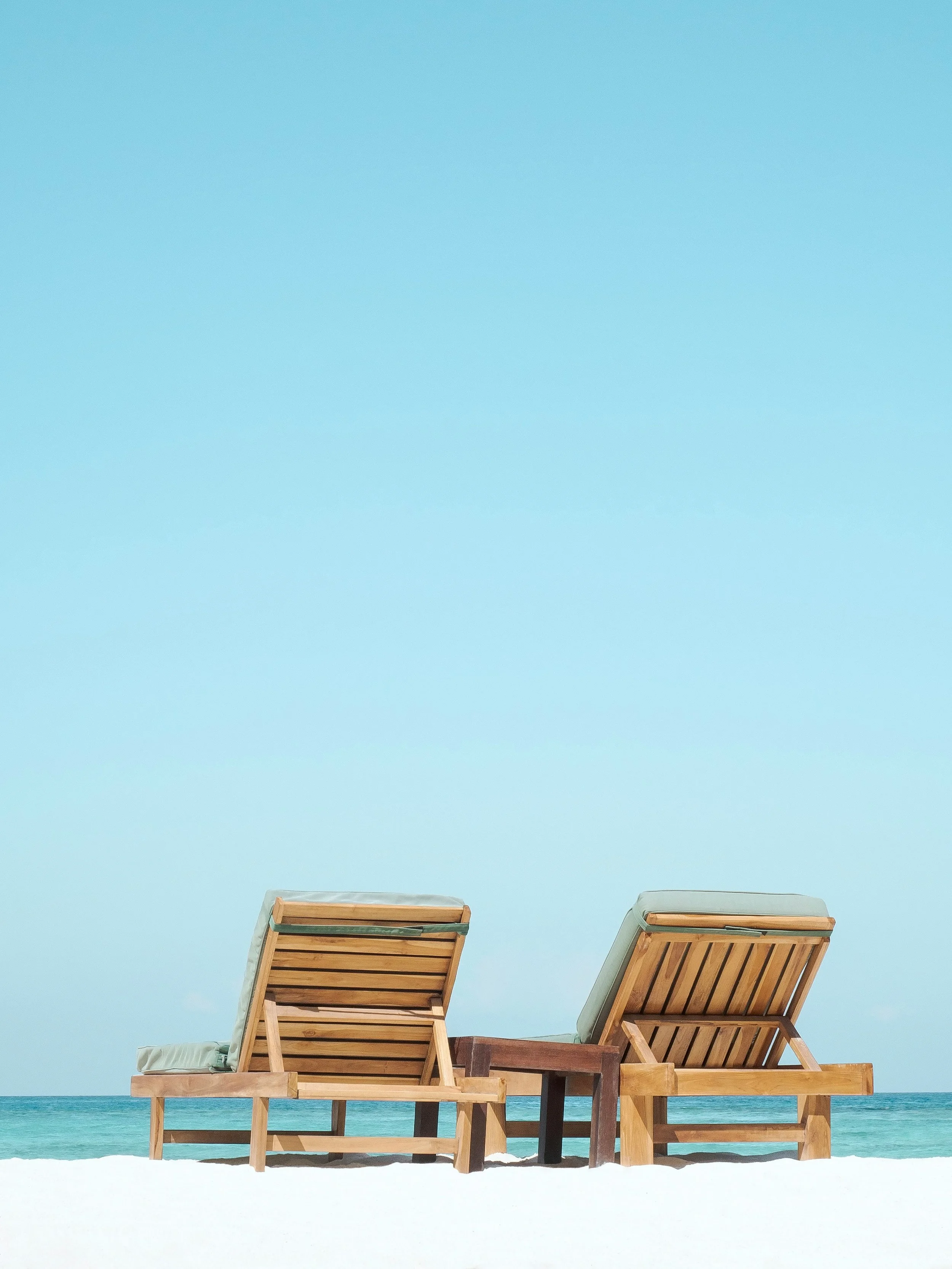Two wooden beach loungers on sand by the ocean under a clear blue sky.