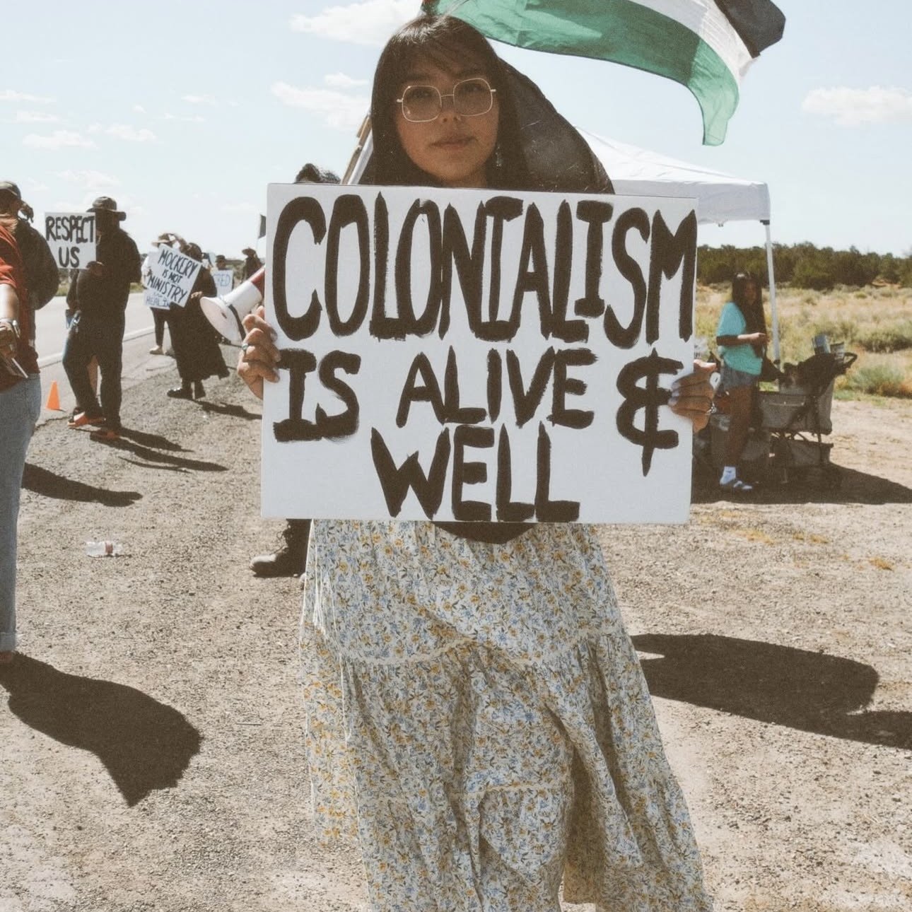 A navajo woman, Louvannina Tsosie, standing outdoors in a protest holding a sign that reads 'Colonialism is alive & well.' She wears glasses and a long floral skirt, with other protesters and signs visible in the background. 