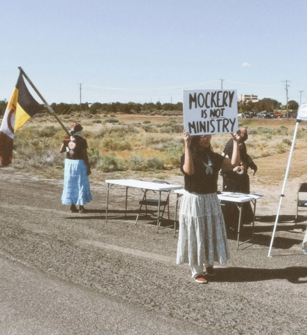 Louvannina Tsosie is holding a sign that reads 'Mockery is not ministry' stands on the side of a road, with another person holding a flag in the background. She advocating for the Diné people.