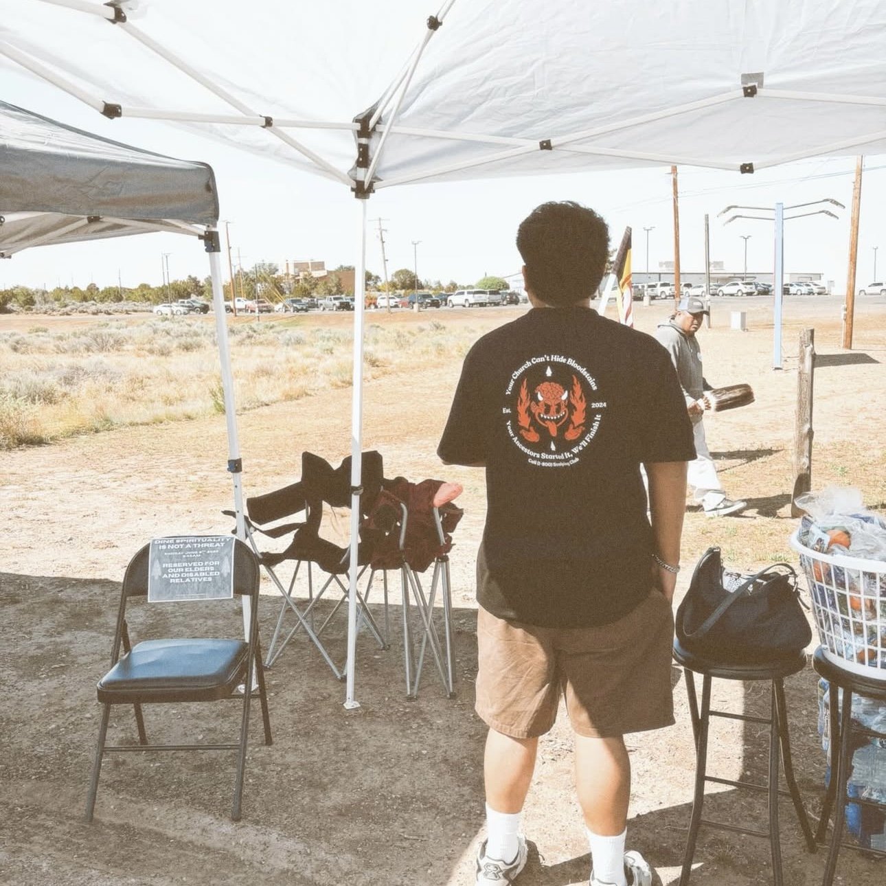 Navajo man with curly hair wearing a black T-shirt with a graphic on the back, khaki shorts, and white shoes, standing under a white canopy in a dirt area with chairs, a basket, and another person in the background.