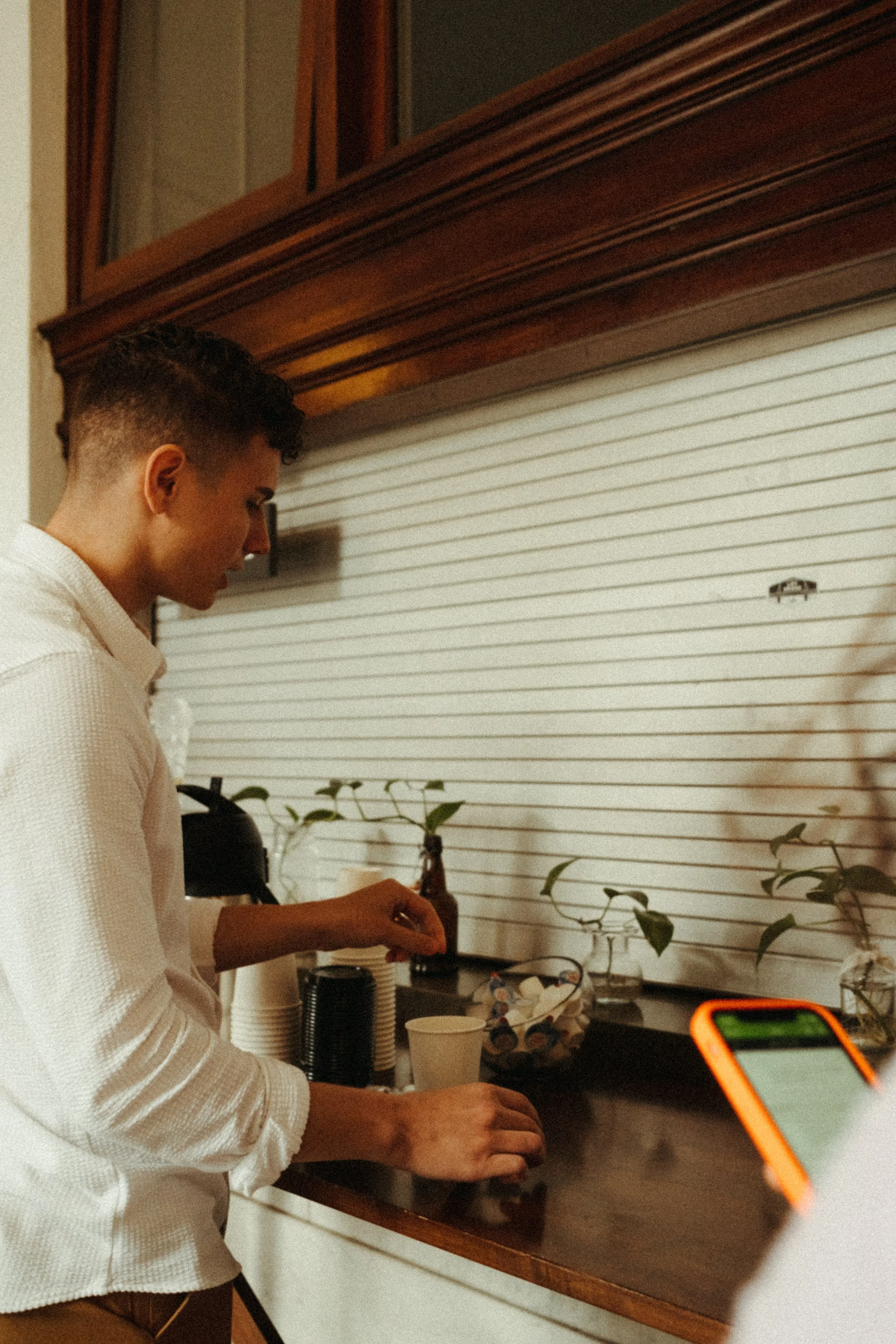 A young man in a white shirt preparing a drink or coffee at a bar counter. The scene includes paper cups, a smartphone with an orange case, and some decorative plants behind him.