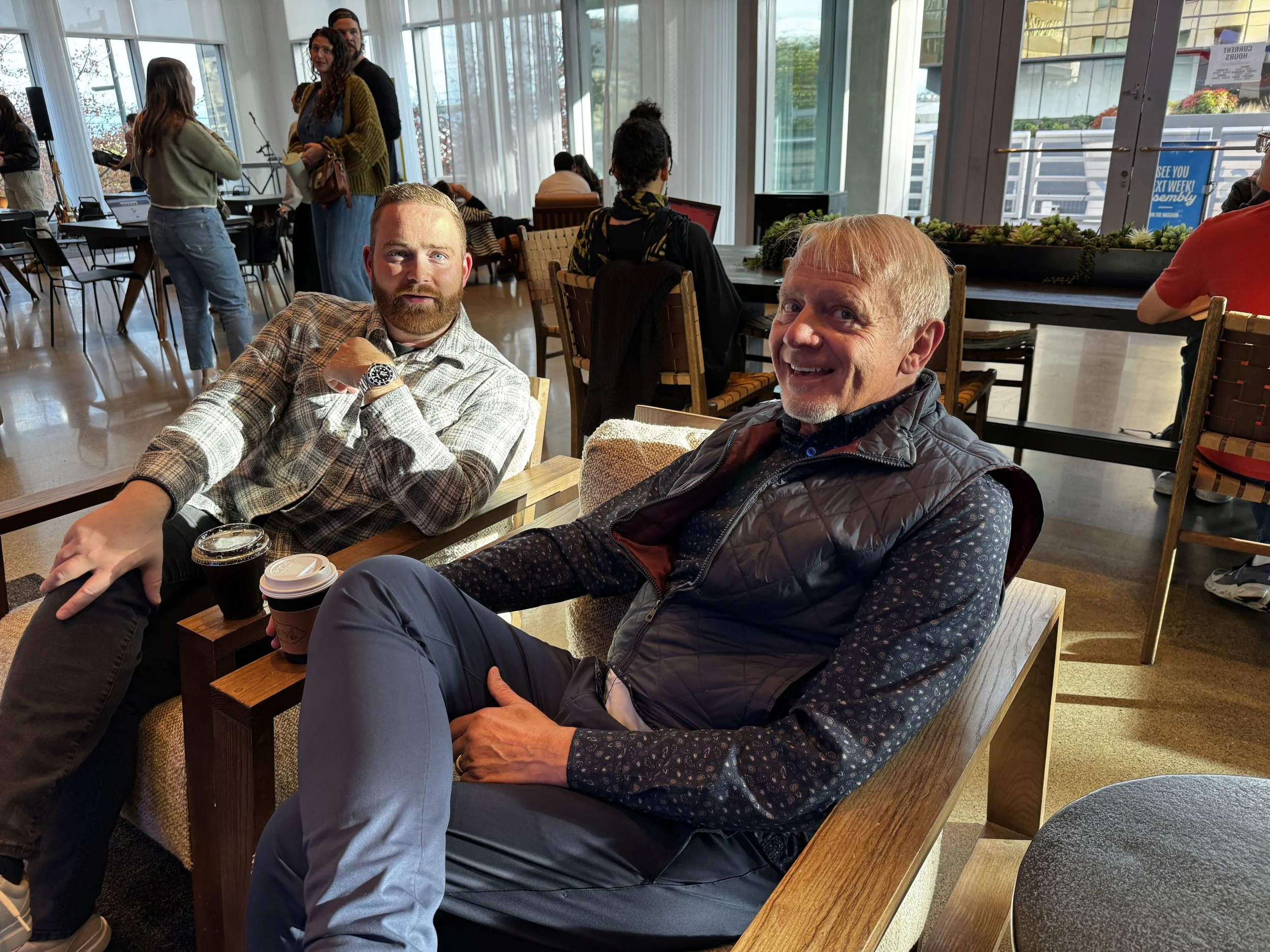 Two men sitting and posing for the photo in a busy coffee shop or cafe with large windows and natural light.