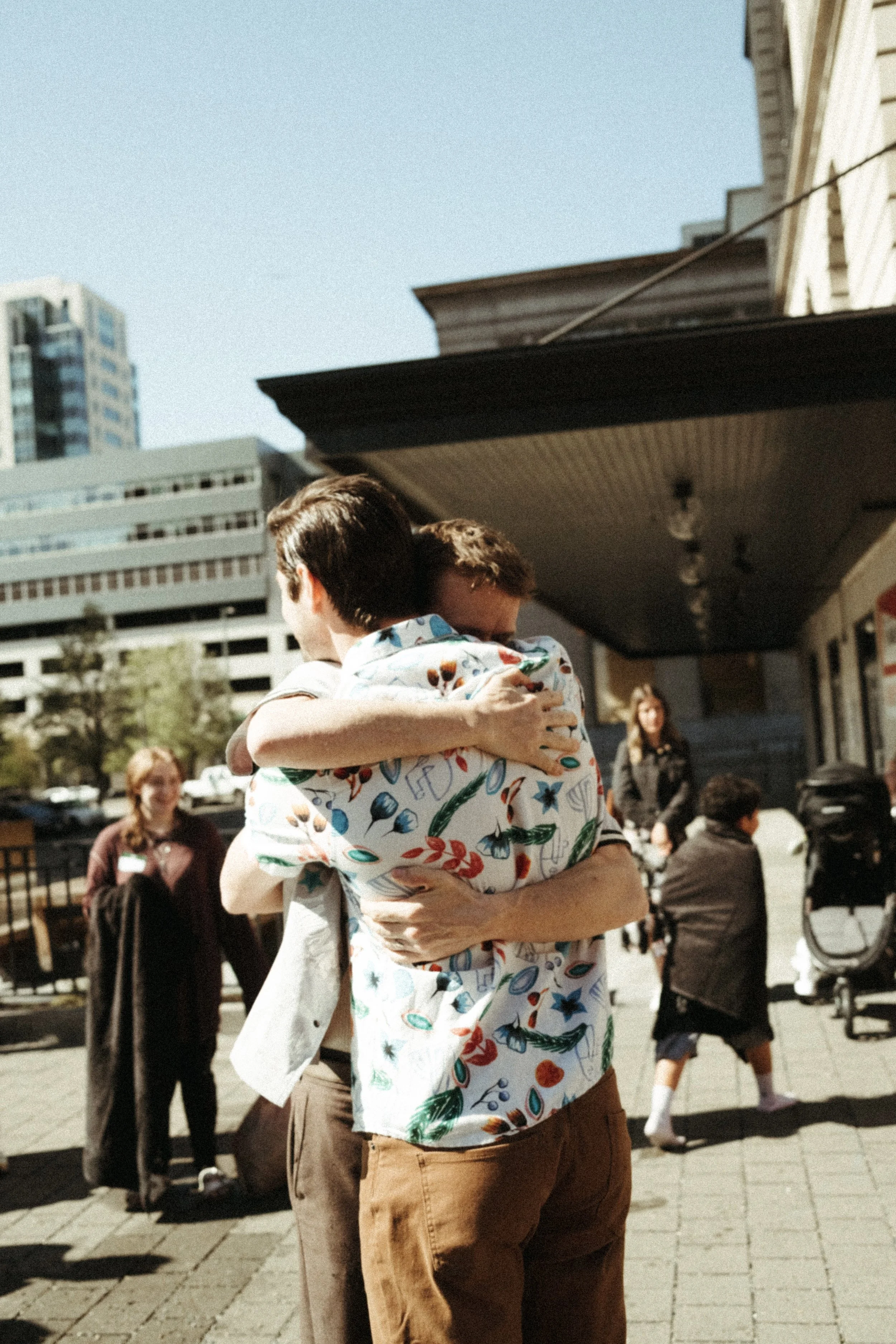 Two men embracing outside on a sunny day, with people and buildings in the background.