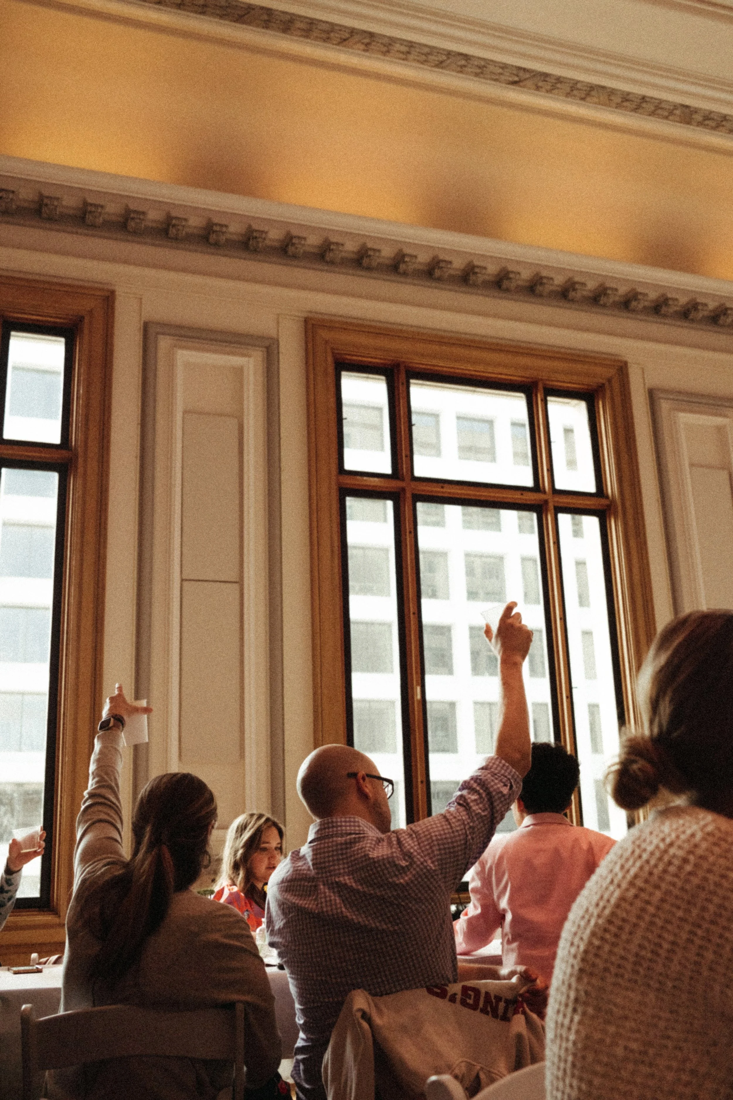 People seated at a table in a room with large windows, some raising their hands during a discussion or presentation.