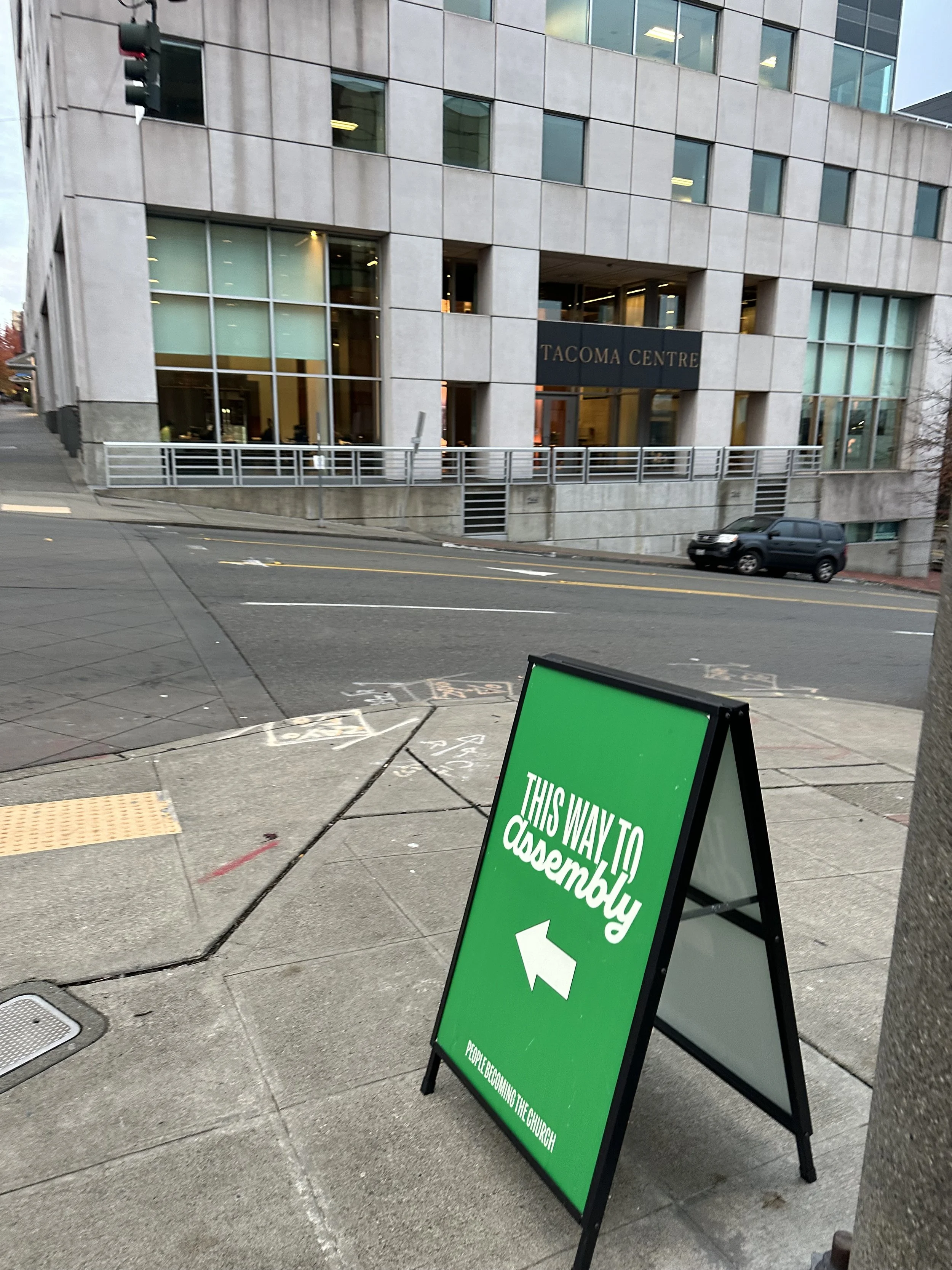 Green sidewalk sign pointing left reads 'This Way to Assembly' with smaller text underneath, located on city sidewalk near a tall building with large windows and a street with parked cars.
