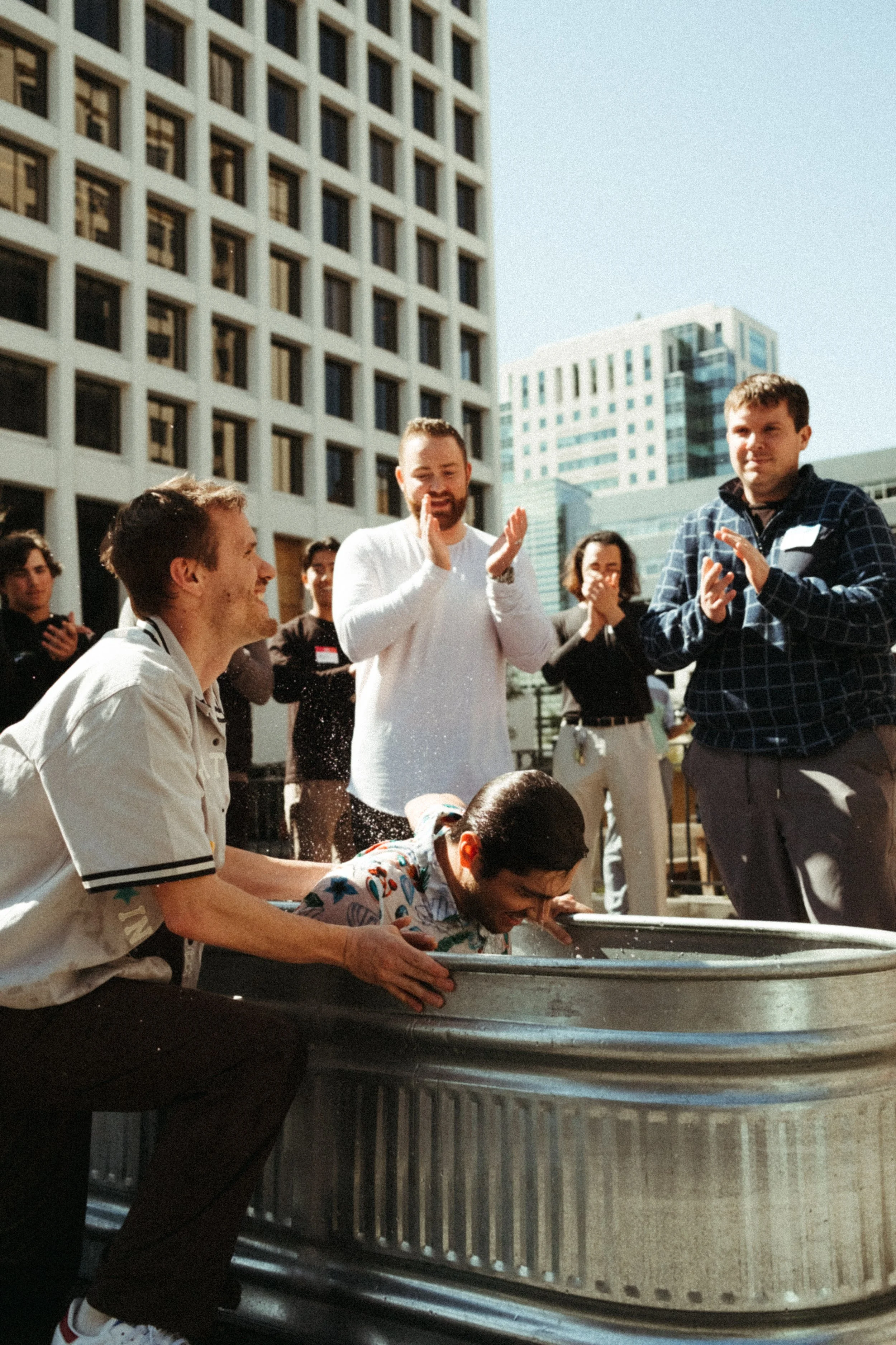 People participating in a water splash challenge outdoors in an urban setting with tall buildings, with one person leaning into a large metal tub while others cheer and applaud.