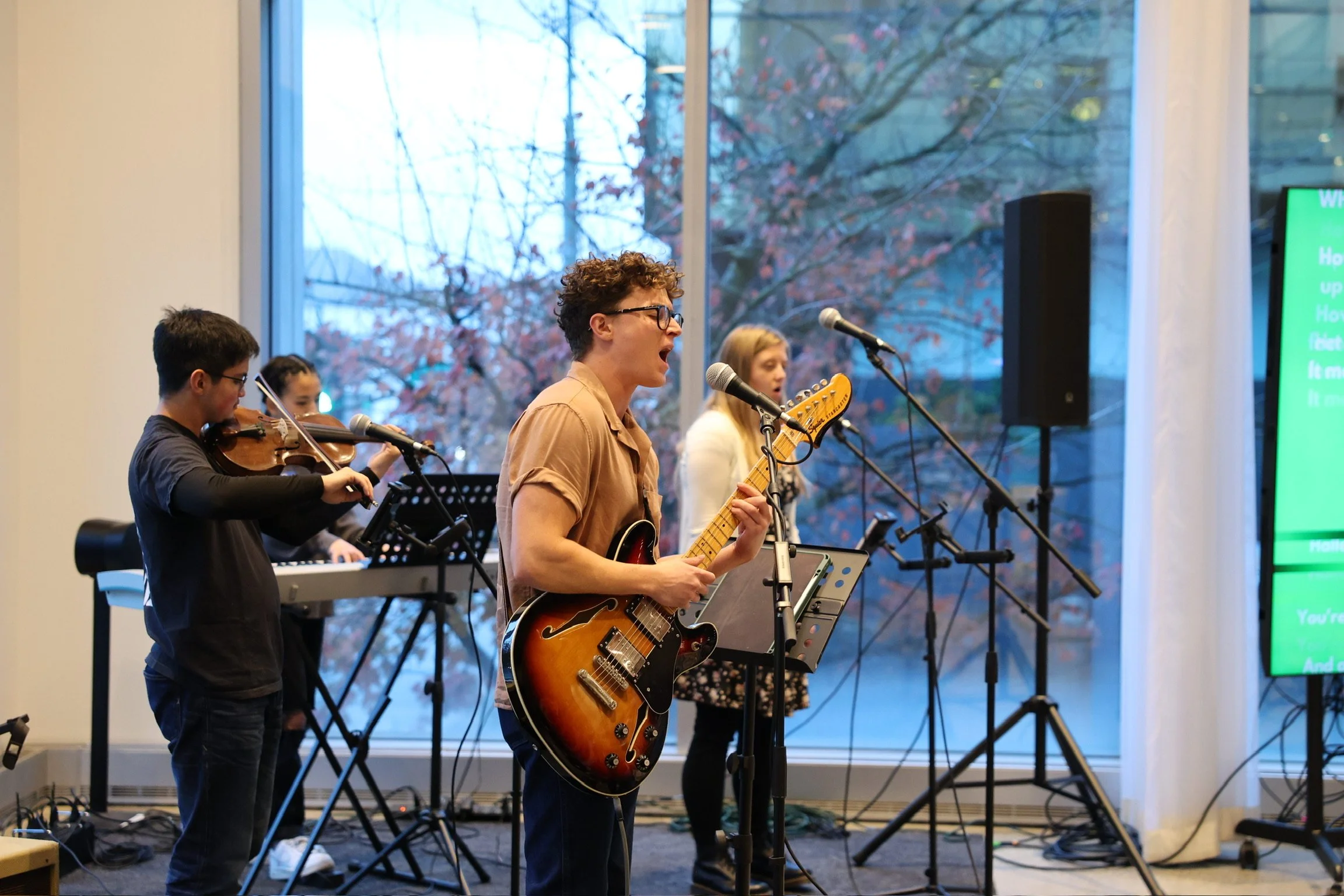 A group of musicians performing indoors near large windows showing trees outside. The lead singer plays an electric guitar and sings into multiple microphones, with two band members playing violin and keyboard in the background.