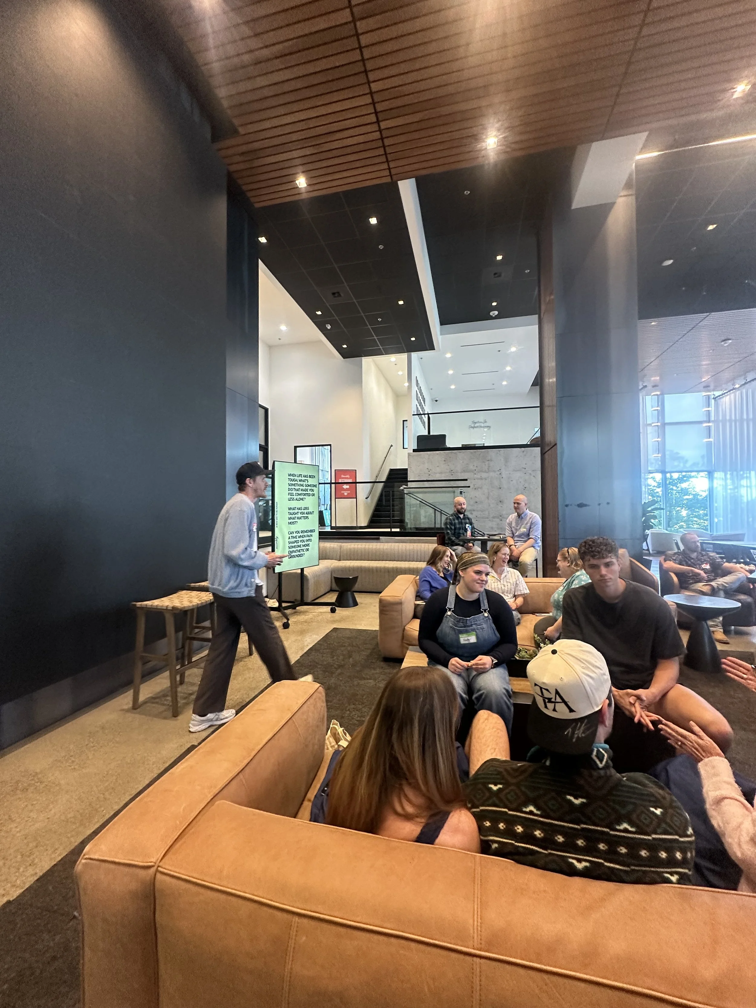 People seated in a modern hotel lobby, listening to a speaker standing next to a display screen.