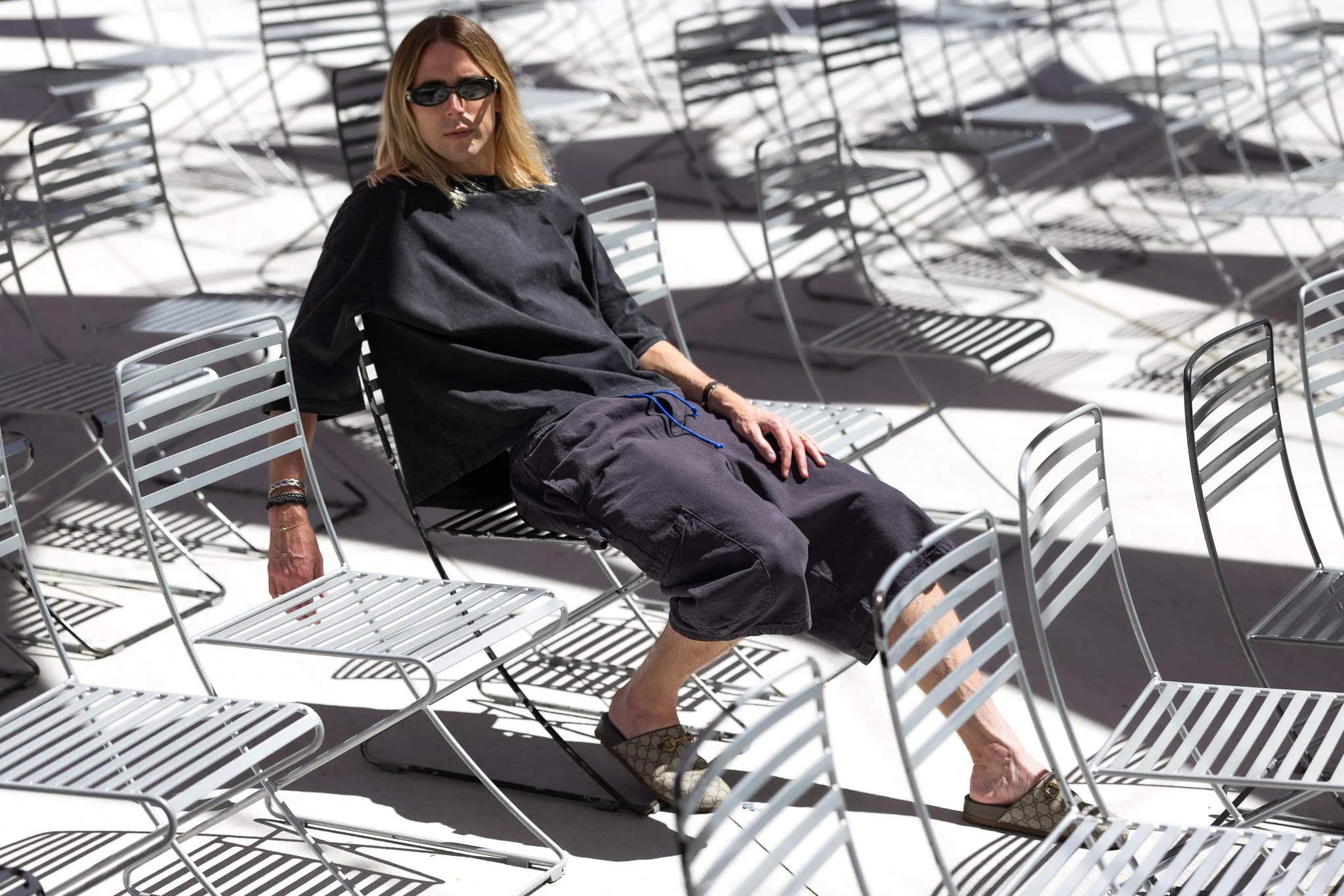 A person with shoulder-length blonde hair wearing sunglasses, a black oversized t-shirt, black shorts, and Gucci shoes is sitting on a metal chair in an area with many empty metal chairs casting shadows on the ground.