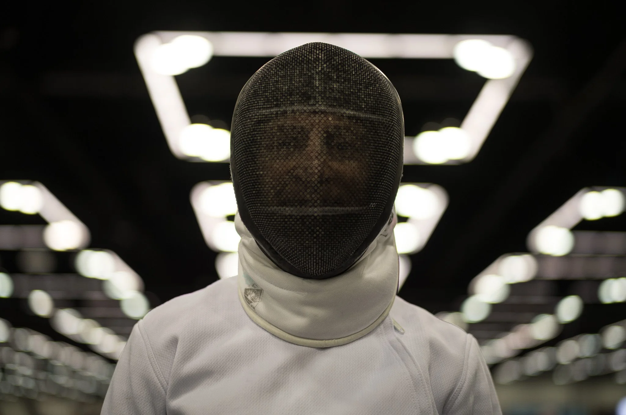 Person dressed in fencing attire with a mask, standing in an indoor space illuminated by overhead lights.
