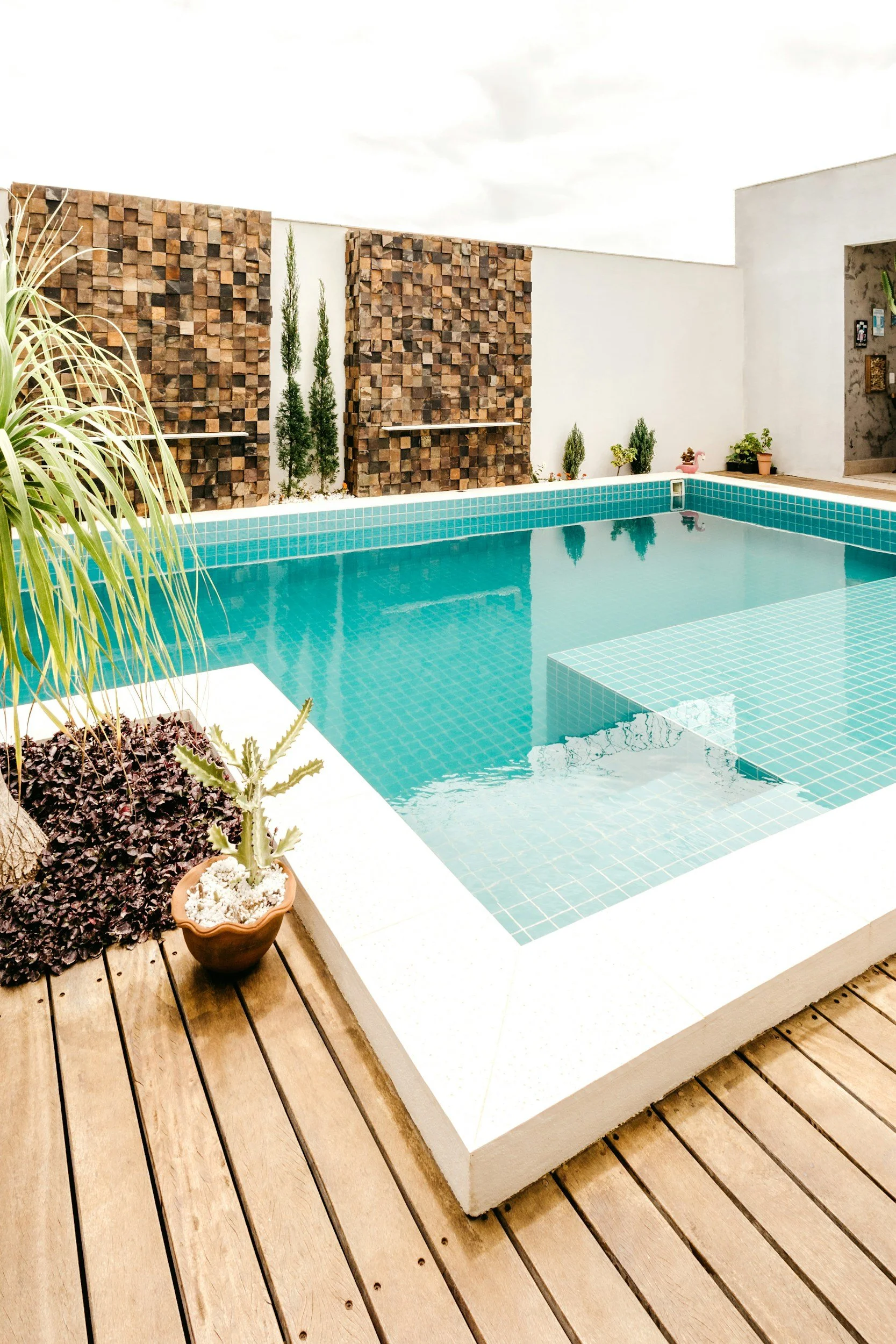 Modern square swimming pool with turquoise tiles, a shallow step area, timber decking, potted plants, and a decorative stone feature wall.