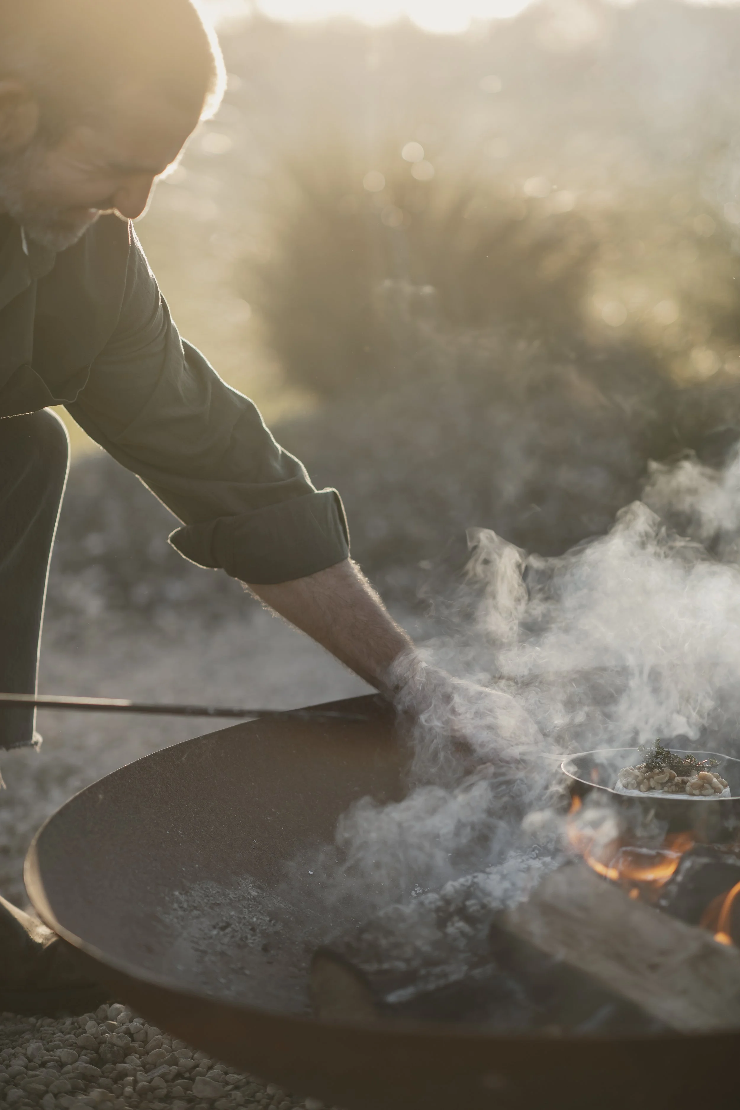 Man cooking over an open flame with smoke rising, wearing a dark jacket and gloves, in an outdoor setting during sunset.