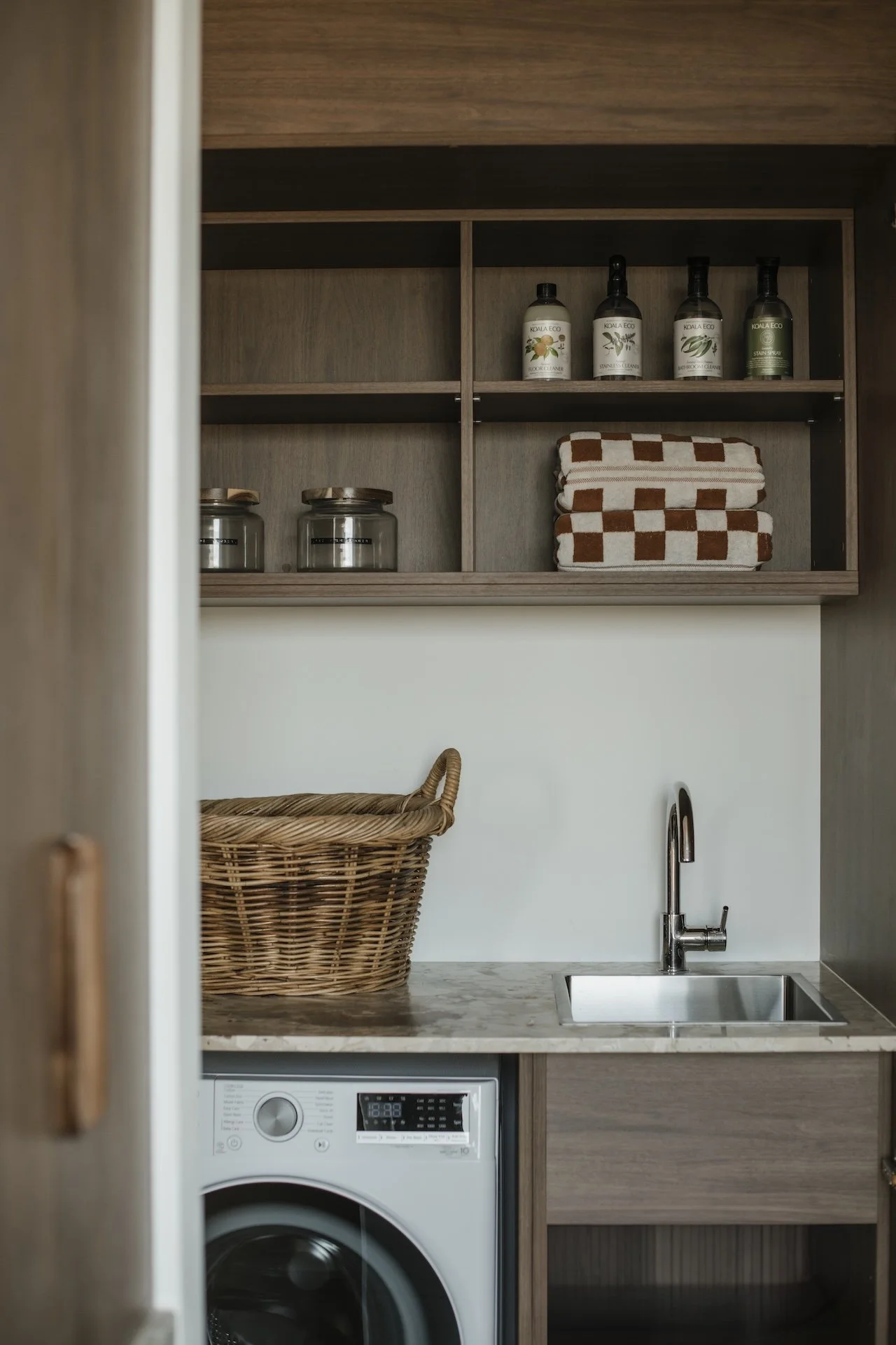 Luxury Interior view of a laundry nook with open wooden shelves containing bottles, jars, and folded towels, a small sink with a faucet, a wicker basket, and a washing machine underneath the marble countertop.