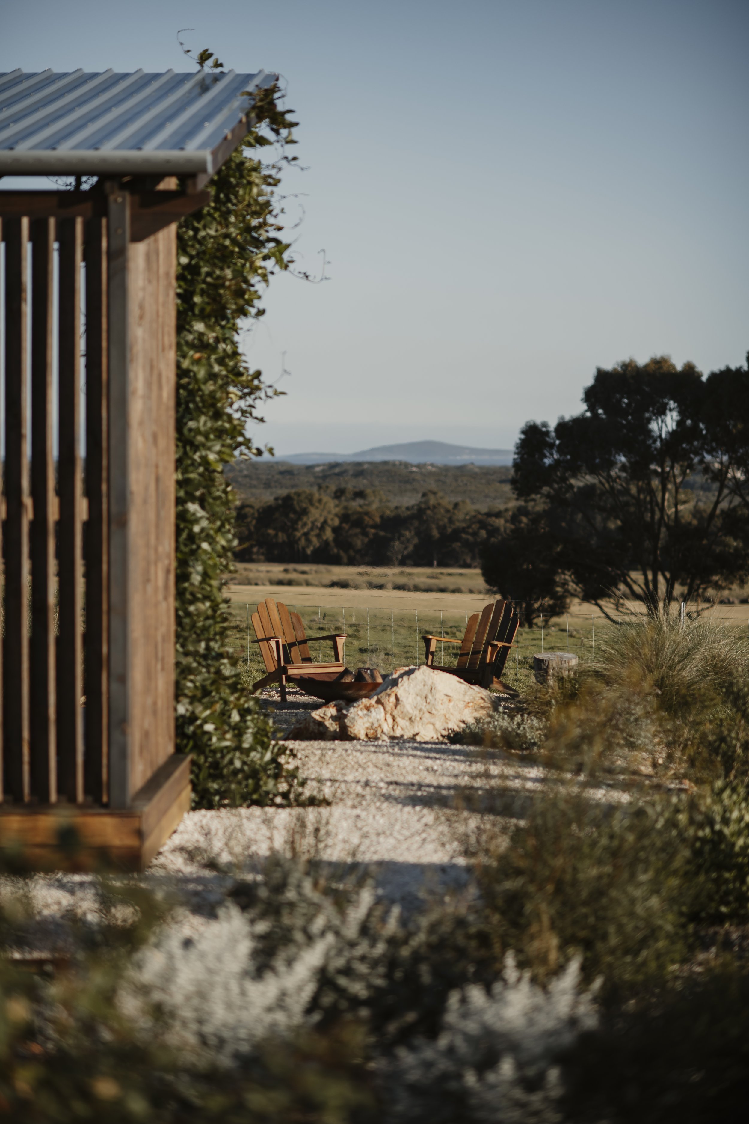 A rustic outdoor seating area with two wooden chairs around a firepit, surrounded by rocks and vegetation, with rolling hills and trees in the background under a clear sky at Box Hill Esperance.