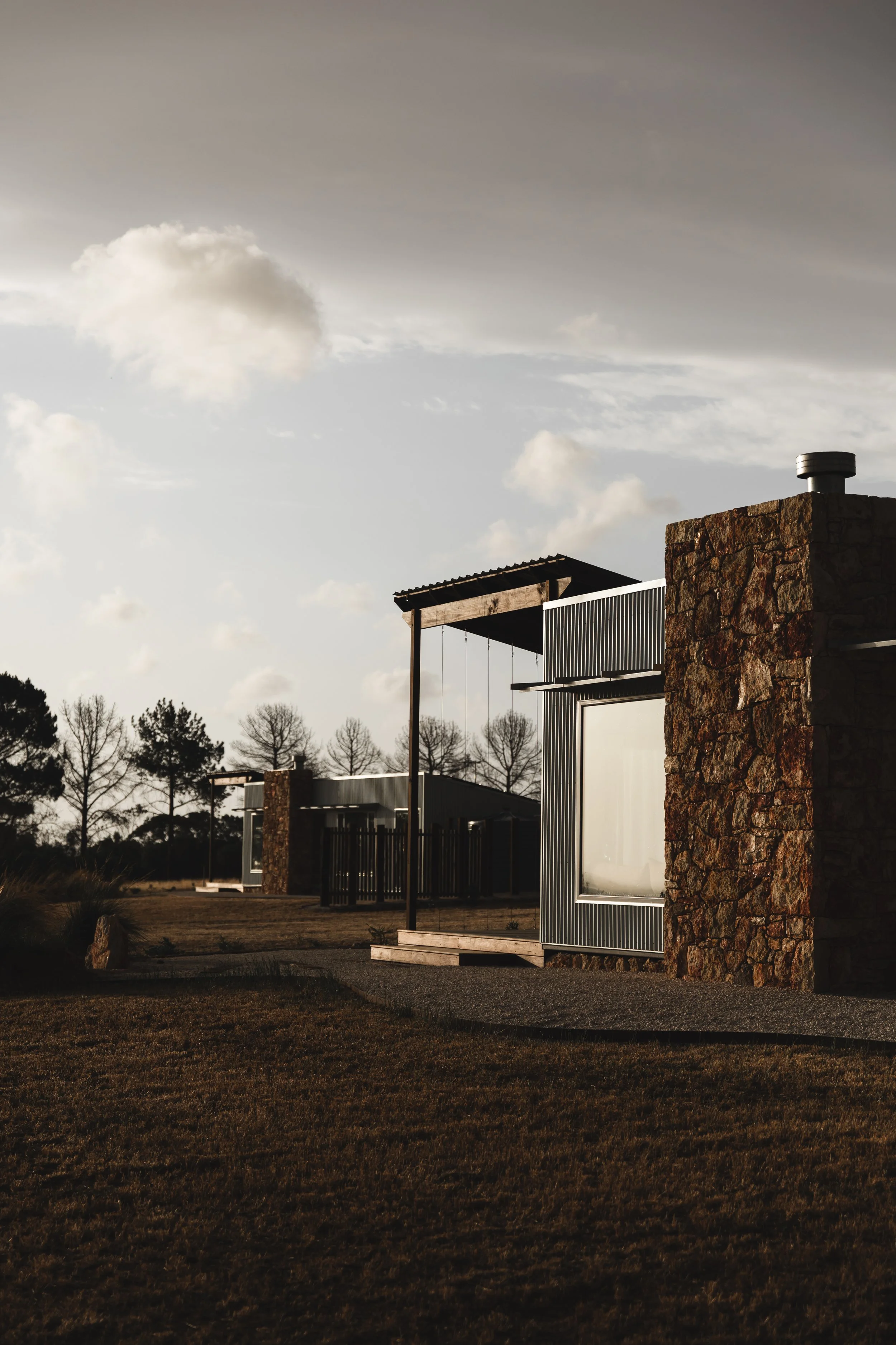 Modern building with stone and metal facade, surrounded by grassy lawn and trees under a cloudy sky at sunset.