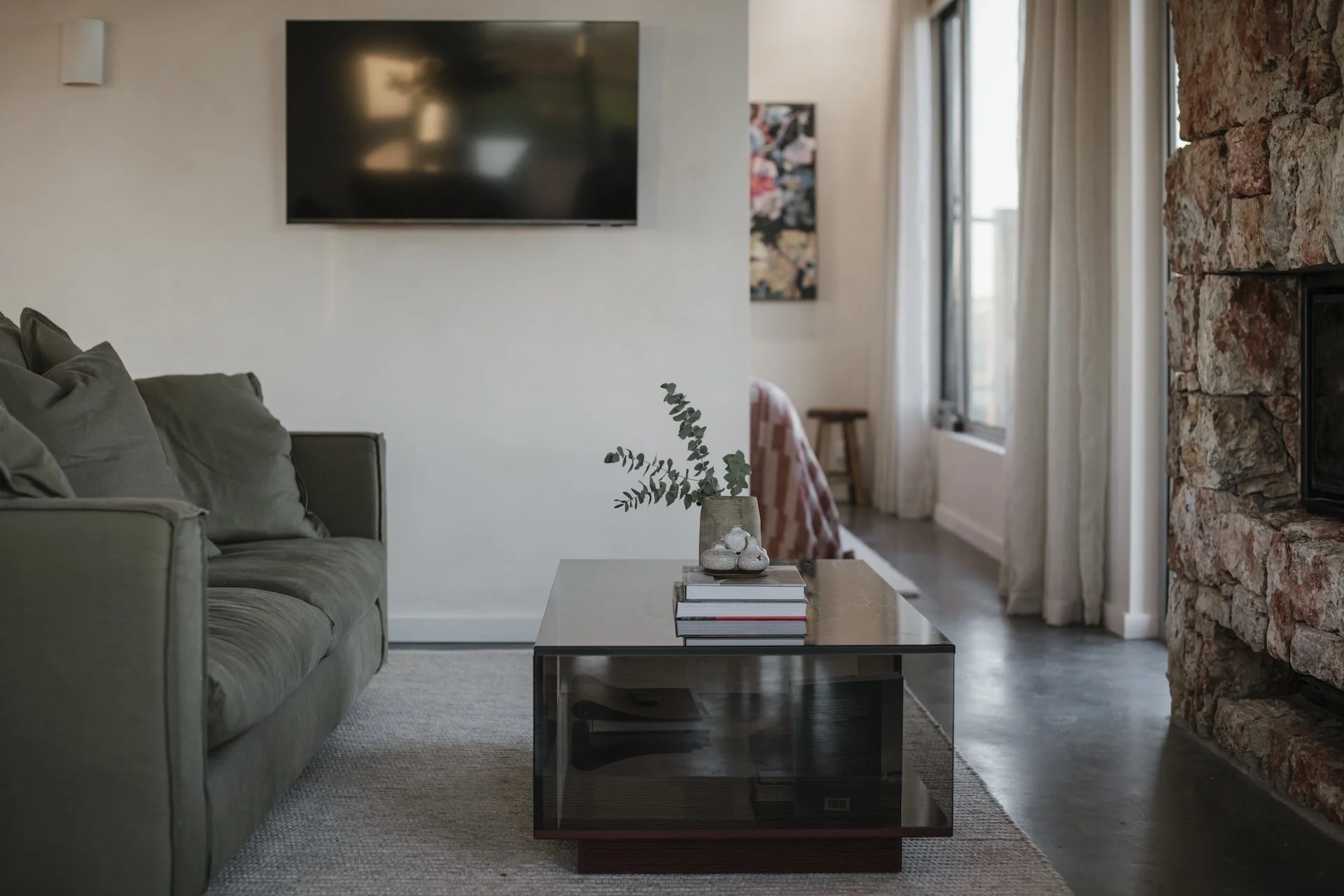 Living room with a green sofa, glass coffee table, and stone fireplace. A wall-mounted TV is above the sofa, and curtains cover large windows in the background.