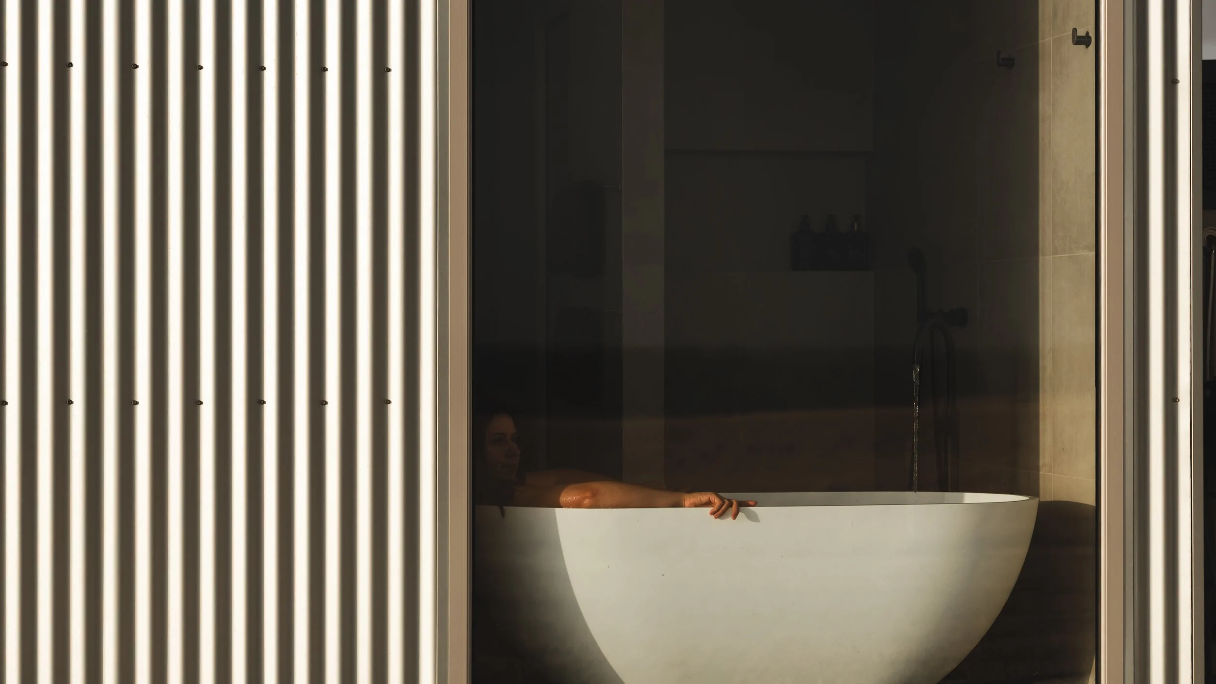 A person relaxing in a bathtub in a modern bathroom at Box Hill Esperance with dark tile walls, visible through vertical metallic slats or beams.