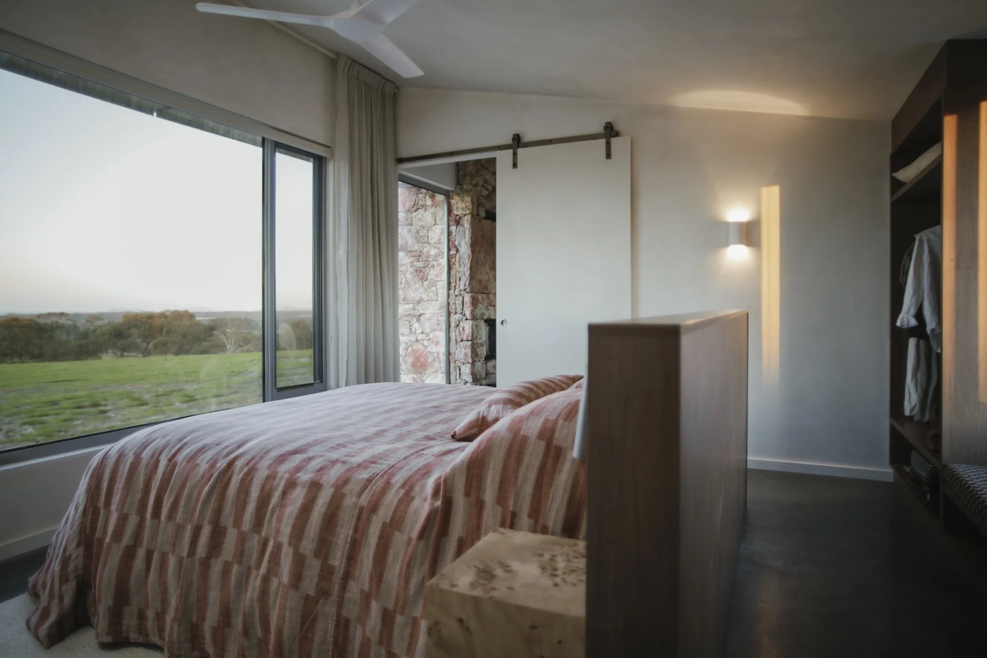 Luxurious modern bedroom with large window showing a green landscape, a bed with striped peach and white bedding, and a sliding barn door leading to an outdoor space.