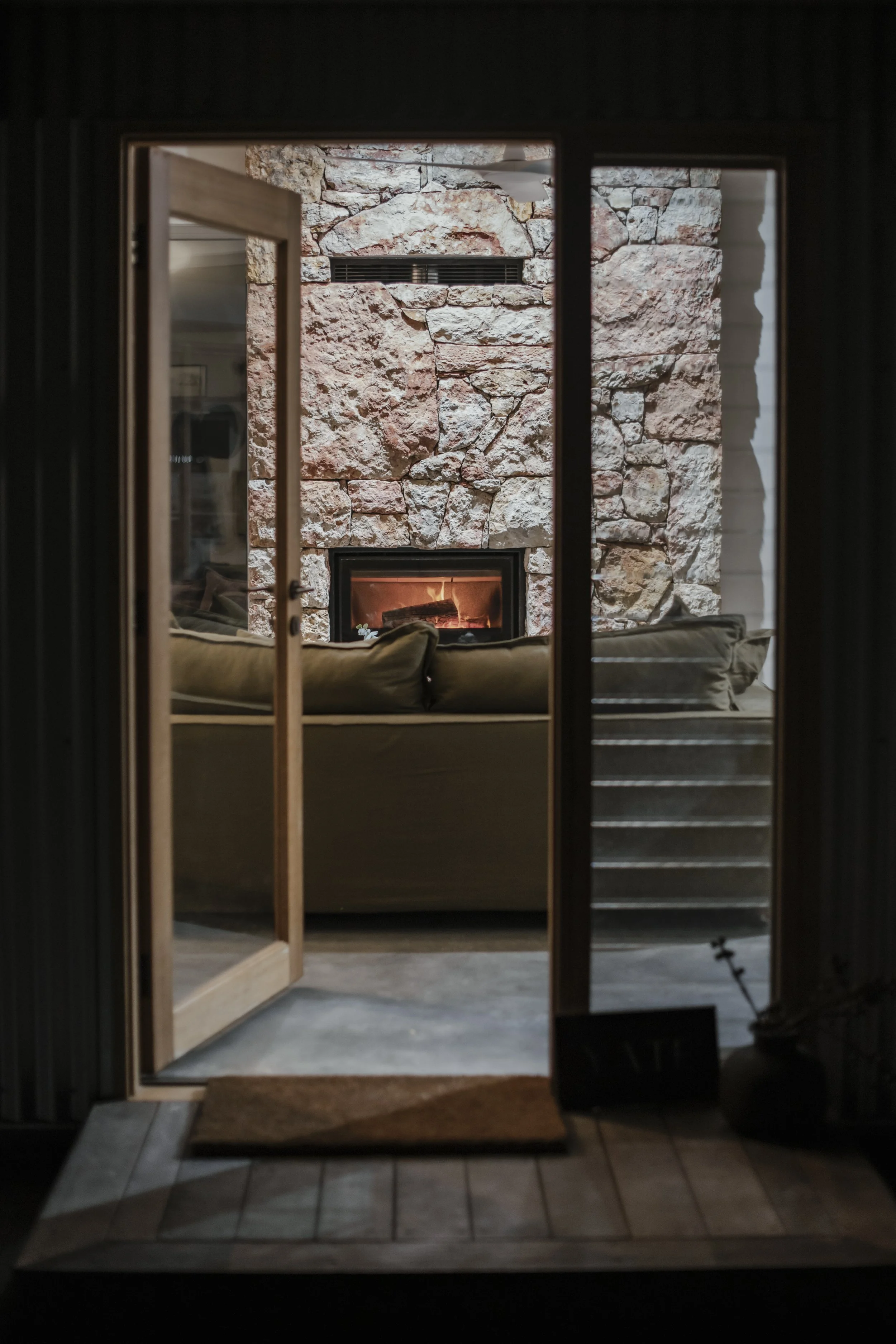 view through a glass door of a living room with a stone fireplace, sofa, cushions, and stairs.