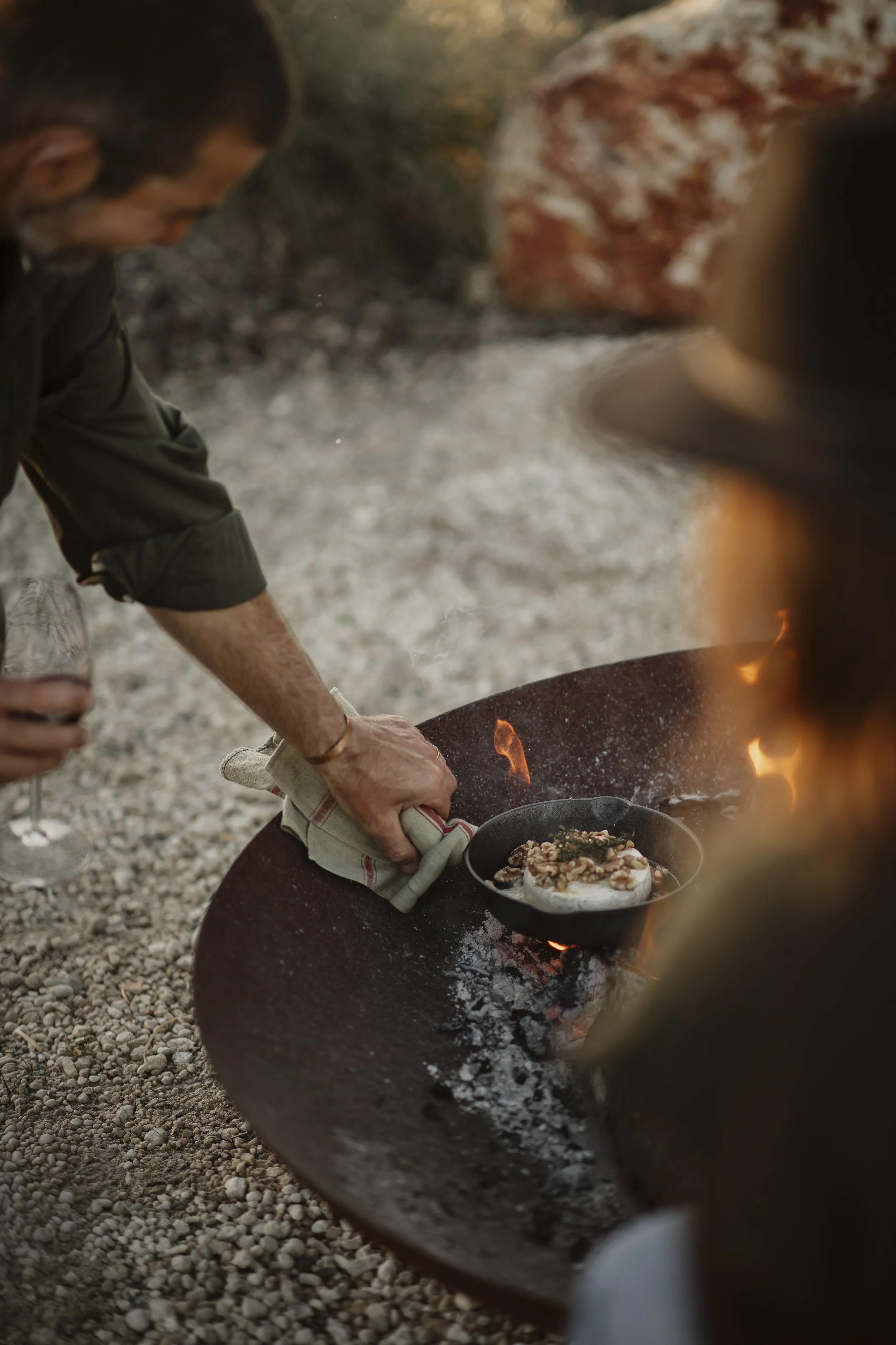 A person cooking food in an outdoor fire pit at Box Hill Esperance, with food in a pan and flames visible.