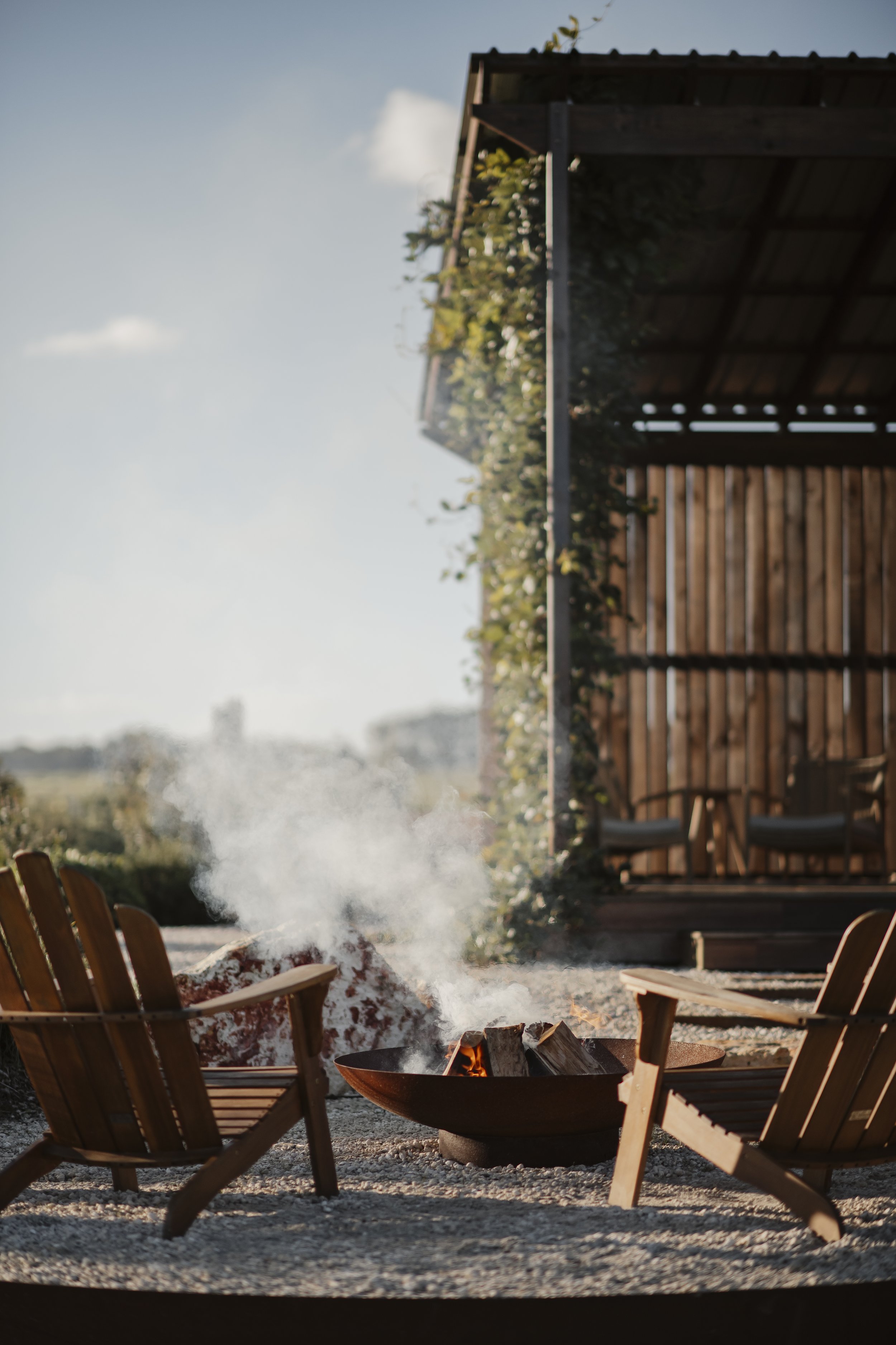 View of luxury cabins at Box Hill Esperance outdoor seating area with wooden chairs surrounding a fire pit, smoke rising from the fire, and a wooden privacy fence in the background under a clear sky.
