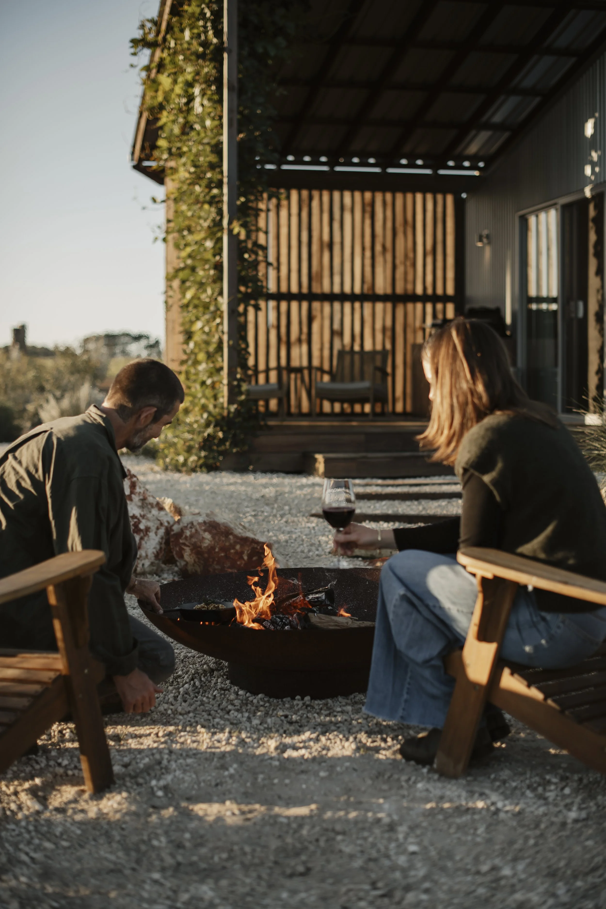 A man and woman sitting around a fire pit on a gravel surface at an outdoor patio, with a wooden fence and house in the background. The woman is holding a glass of red wine, and both are relaxing in chairs.