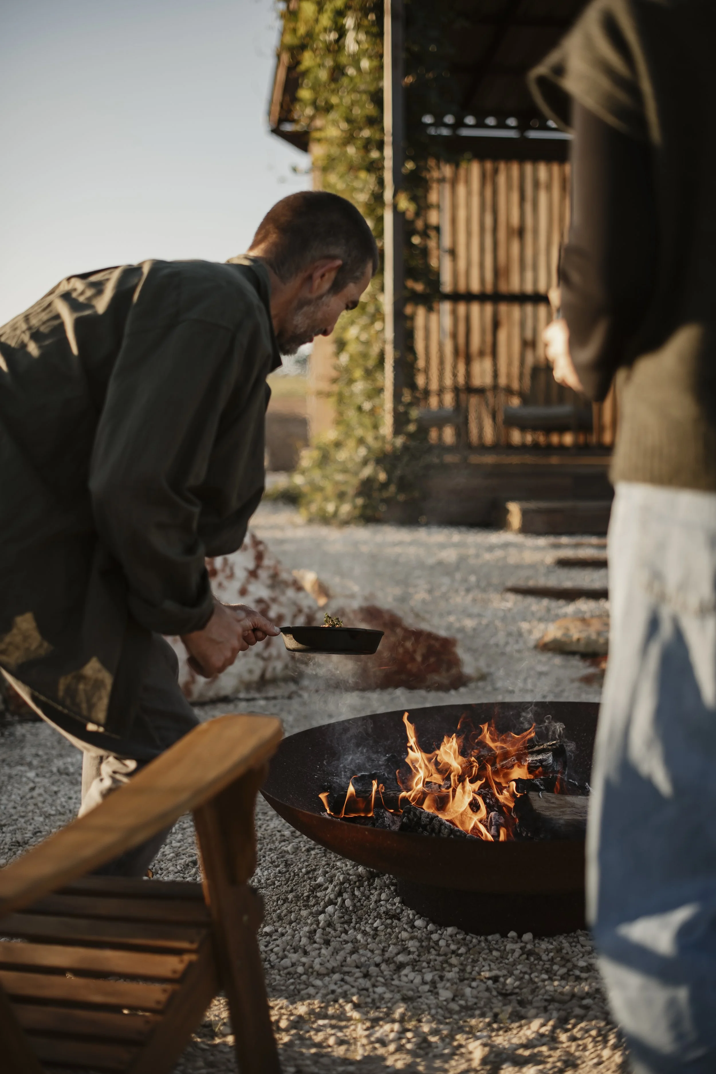 People gathered around a fire pit outdoors during sunset, some cooking or tending to the fire, with a wooden structure and trees in the background.
