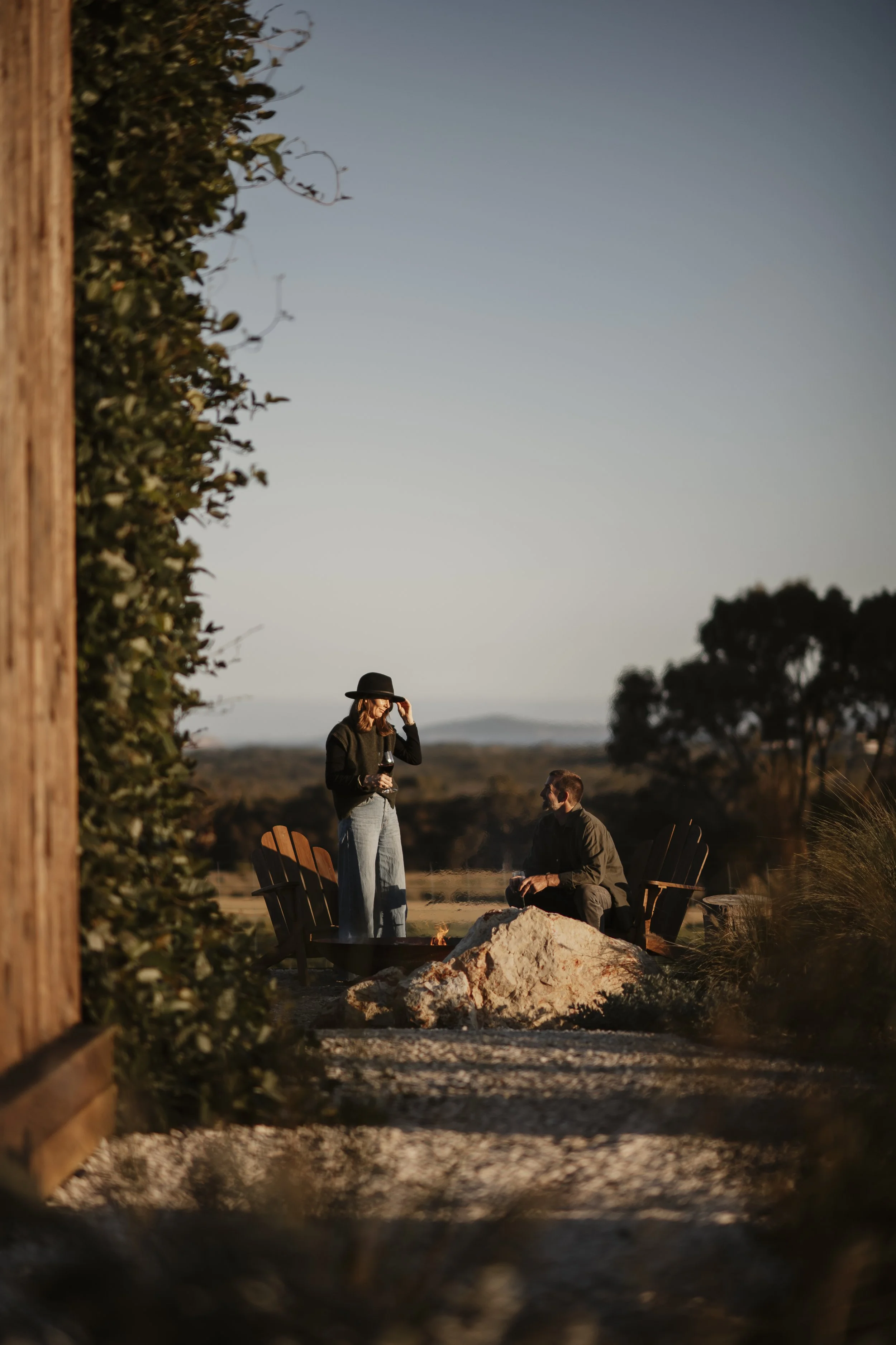 Three people outside at sunset, with one woman standing holding a glass of wine, and a man sitting near a fire pit. They are surrounded by Adirondack chairs and rocks.